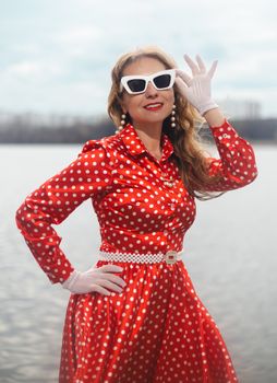 Stylish woman in a vintage red polka dot dress posing by a lake, exuding classic elegance and glamour.