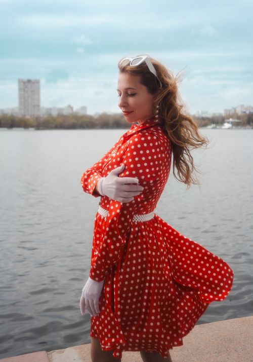 Free Stylish woman in vintage red polka dot dress by a serene waterfront setting. Stock Photo