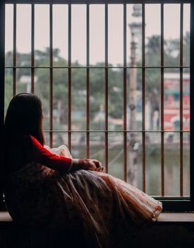A woman in traditional attire gazes out a window with a contemplative look in an indoor setting.