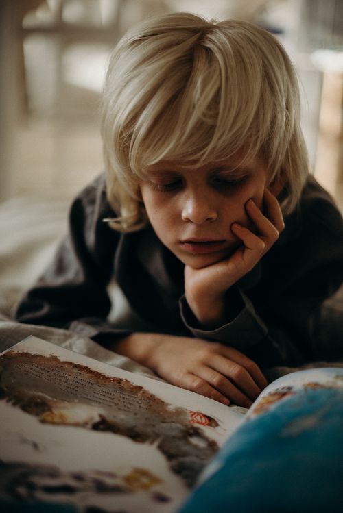 Free A young boy with blonde hair reads a book indoors, deeply focused and thoughtful. Stock Photo