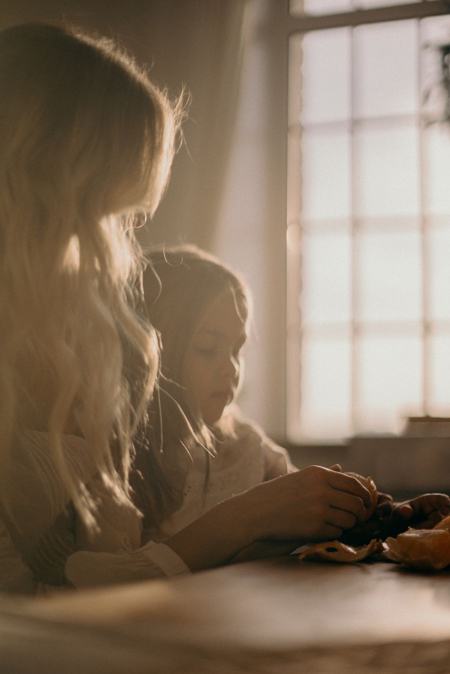 A mother and daughter sitting close together beside a sunlit window at home.