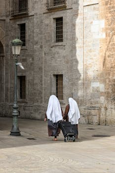 Two nuns walking with a stroller by historical buildings in Valencia, Spain, showcasing serene urban life.