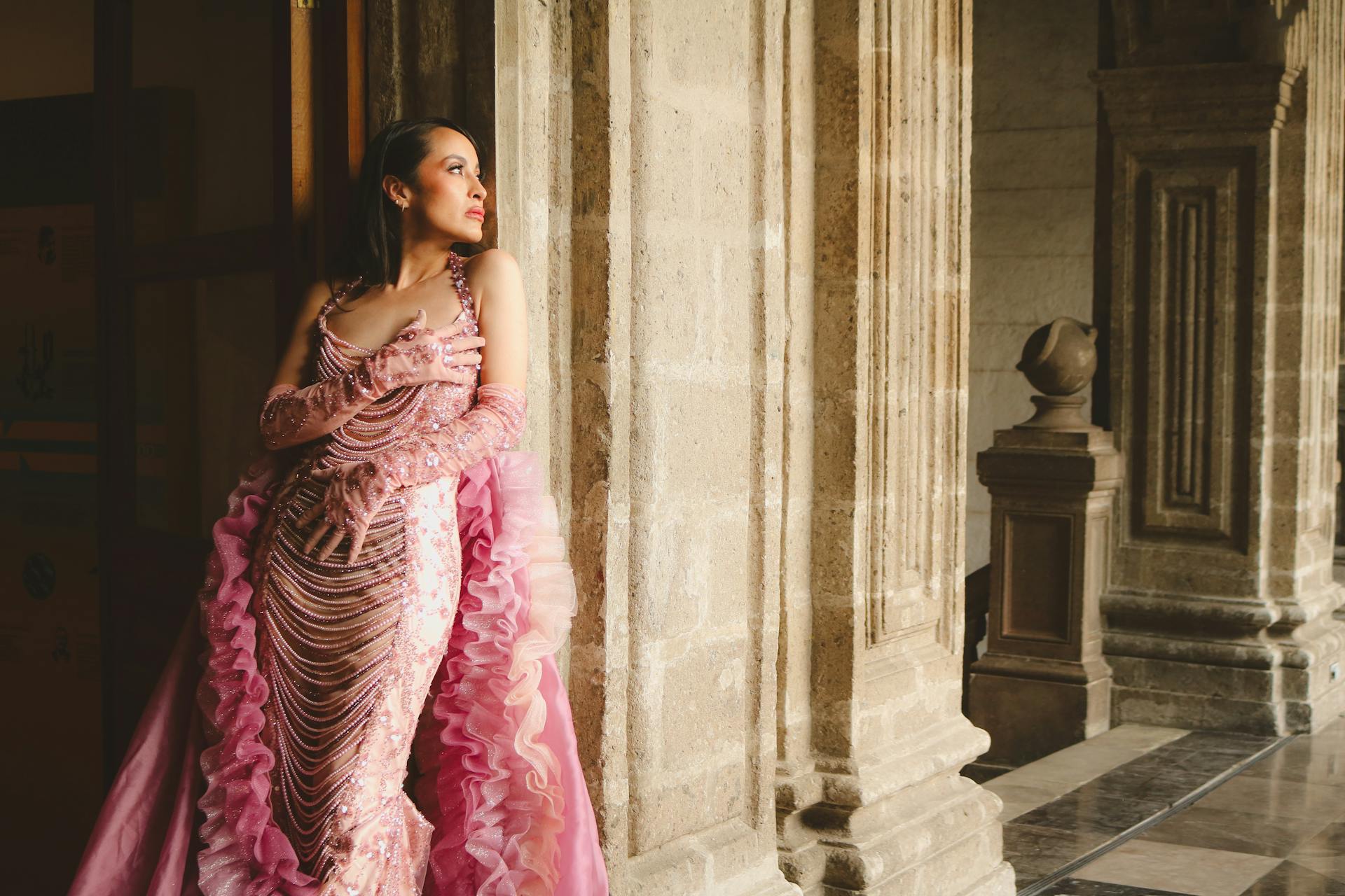 Woman in ornate pink gown leaning against stone pillars in a historic setting.