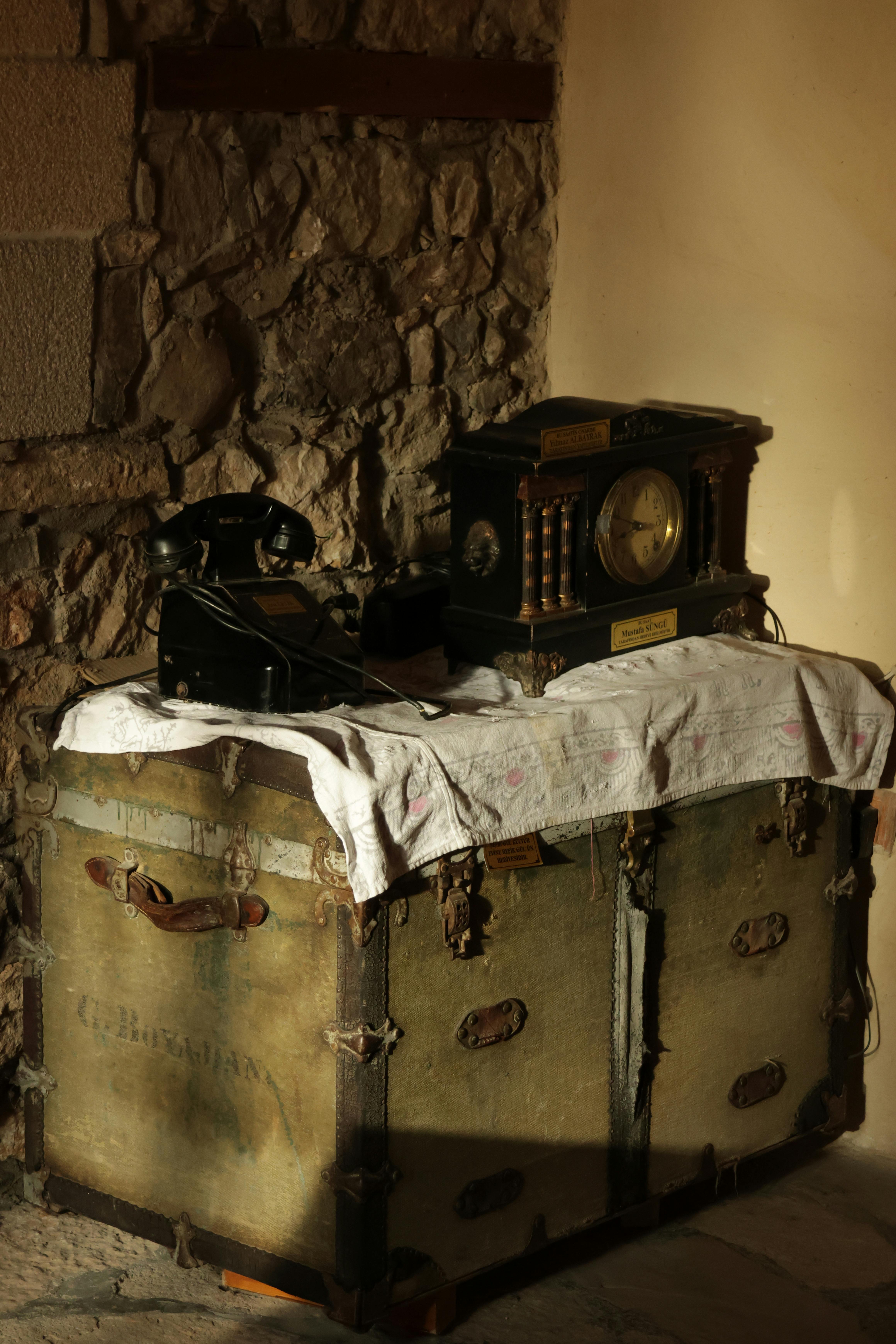 A vintage setup featuring an antique phone and clock on a rustic trunk indoors.