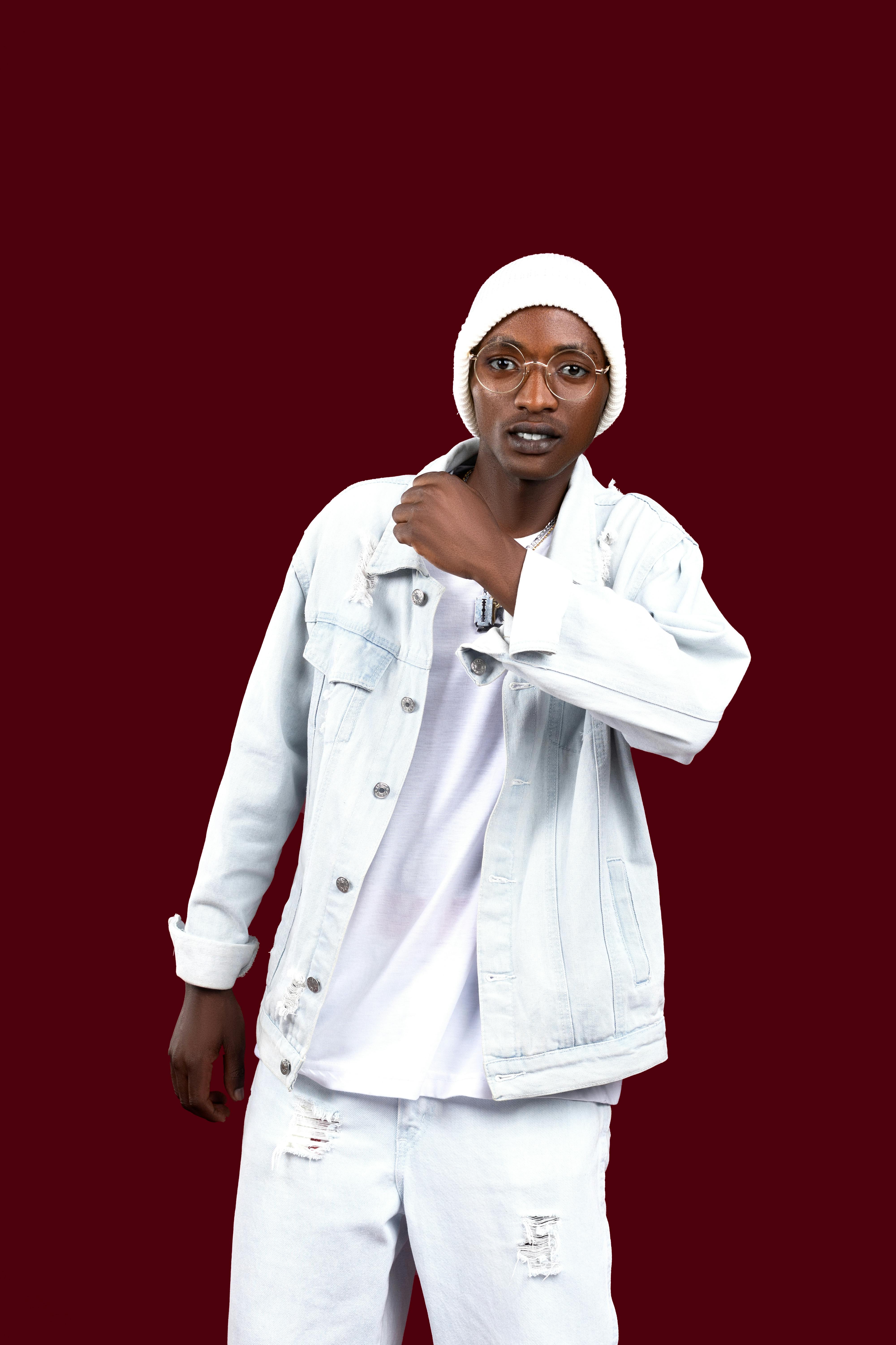 A fashionable young adult in white denim and beanie against a red background.