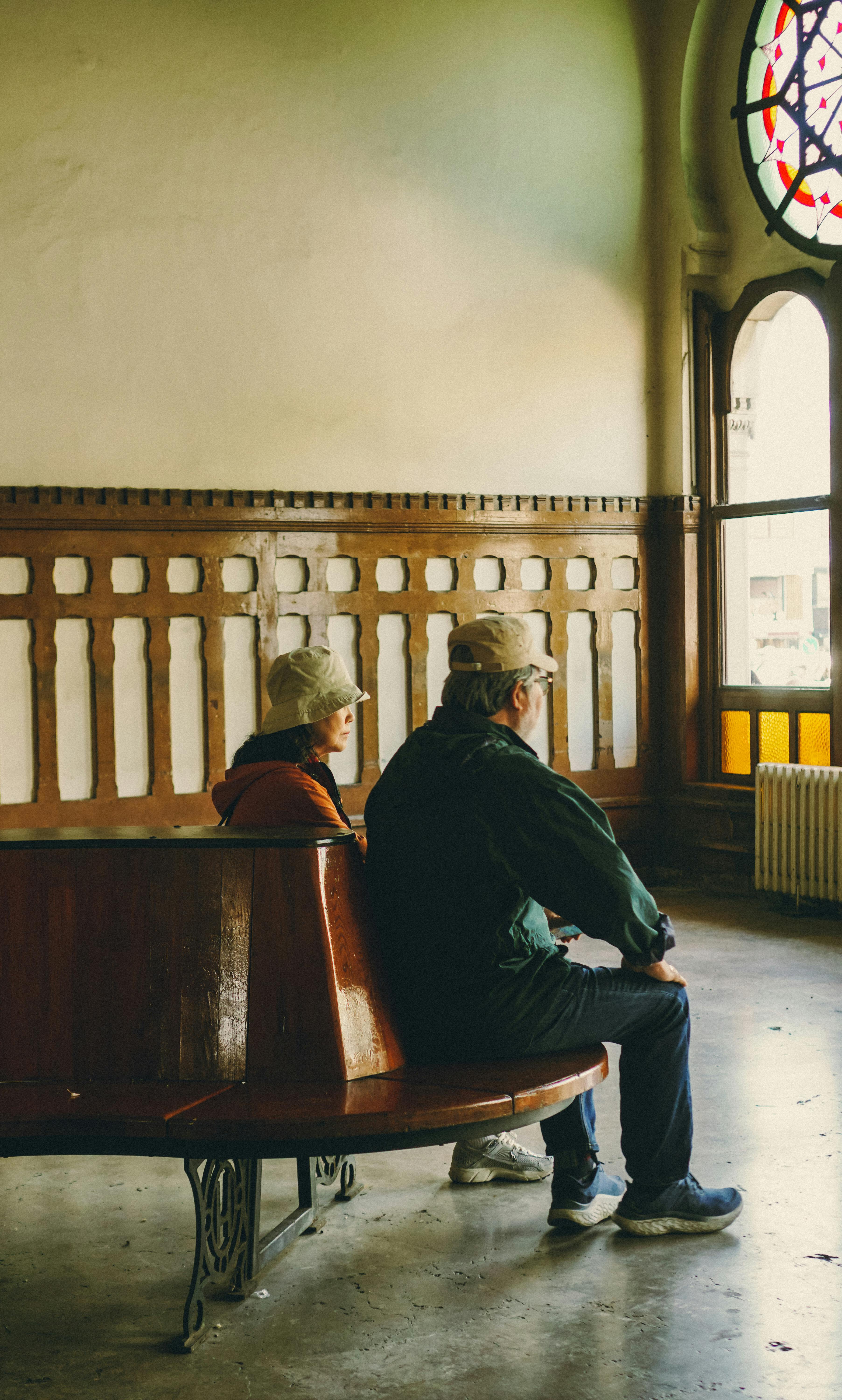 Two seniors sitting on a wooden bench inside a vintage train station, gazing outside.