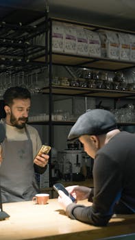 A barista and customer interact at a cafe counter, both engaged with smartphones.
