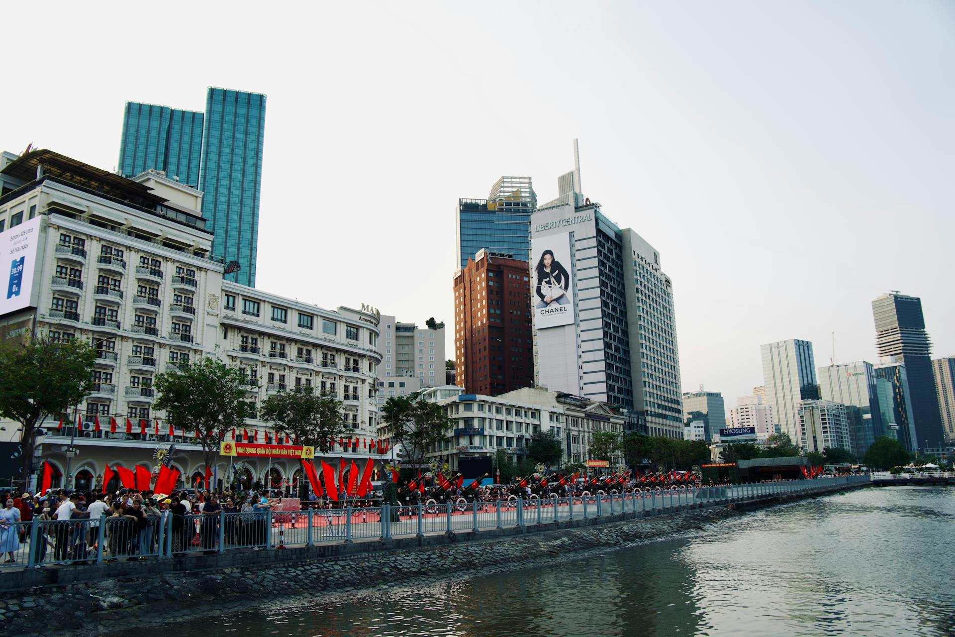 A bustling cityscape along a riverside with high-rise buildings and vibrant flags.