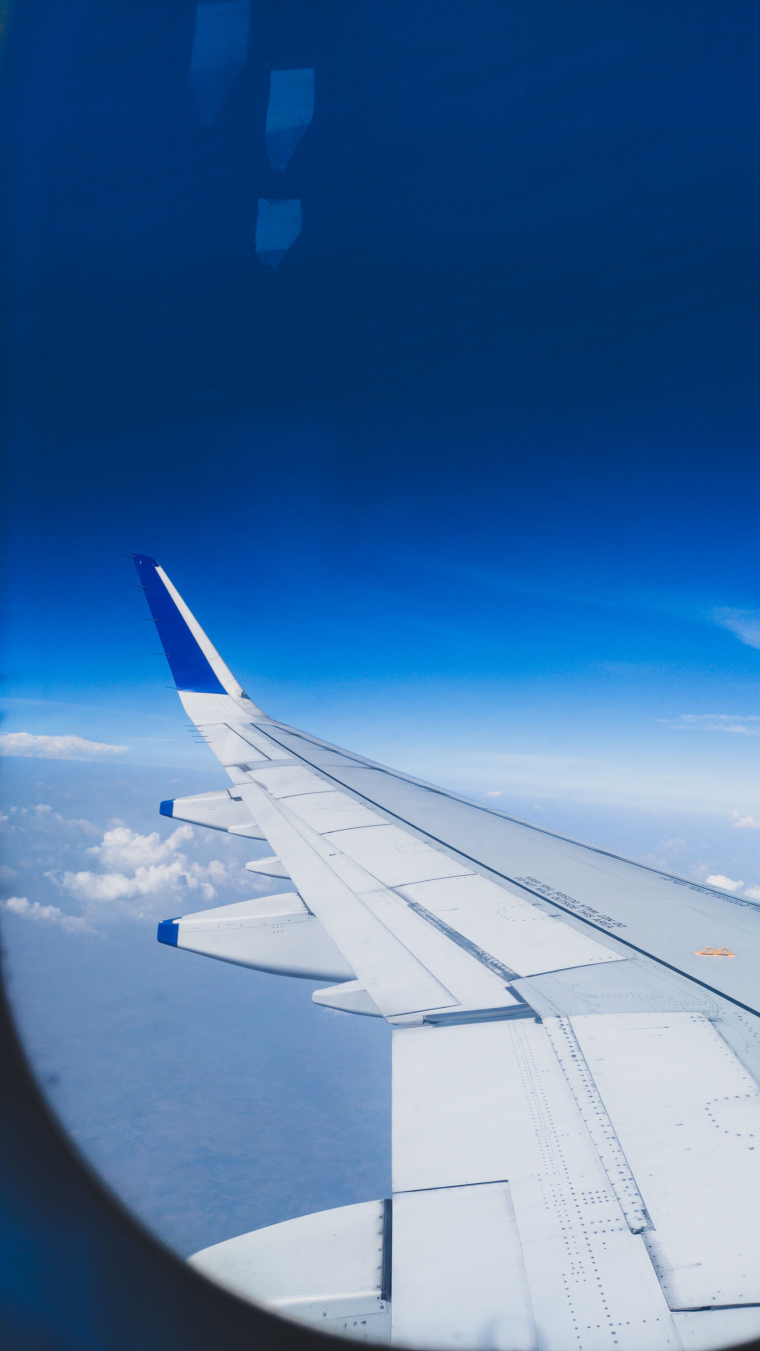 Fotomural Un ala de avión vista desde una ventana contra un cielo azul claro con nubes, capturando la esencia del viaje.