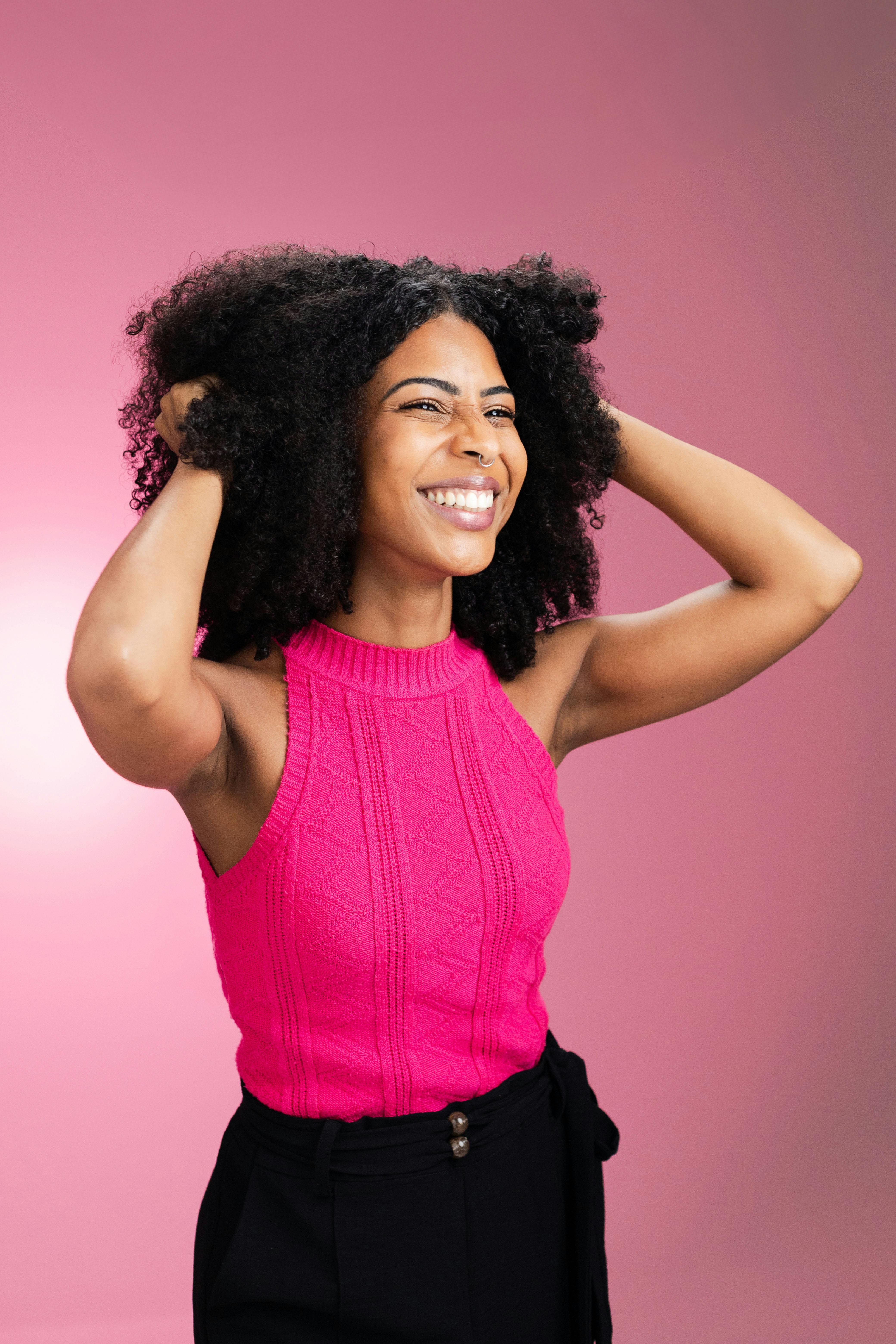 Smiling woman with curly hair wearing pink sweater, expressing joy against a pink background.