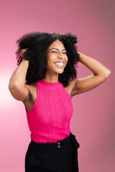 Smiling woman with curly hair wearing pink sweater, expressing joy against a pink background.
