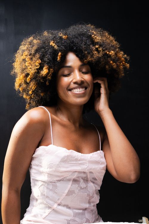 Free A joyful woman with voluminous curly hair smiling in a pink dress against a dark background. Stock Photo