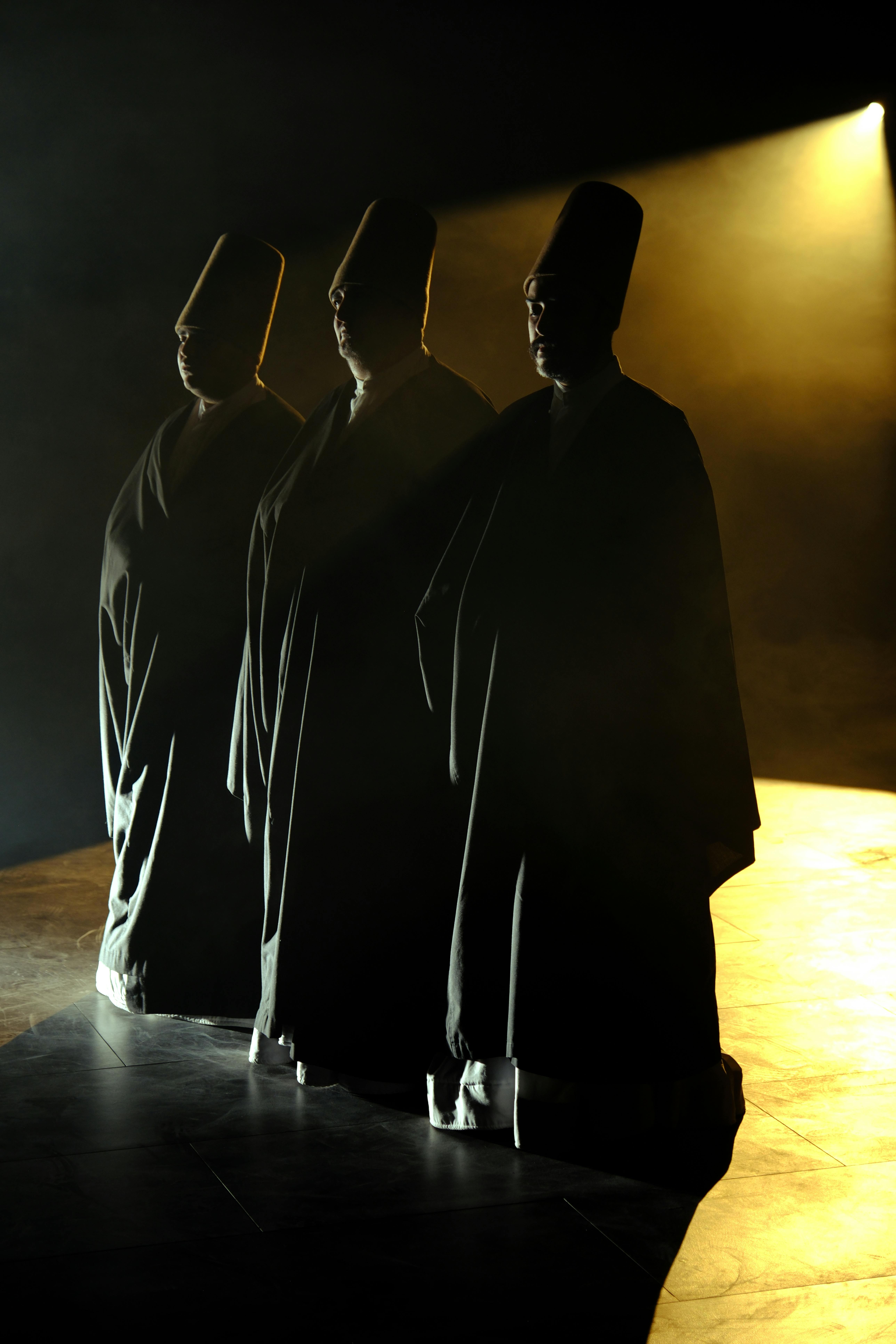 Three whirling dervishes stand illuminated by soft spotlight, creating a mystical ambiance.