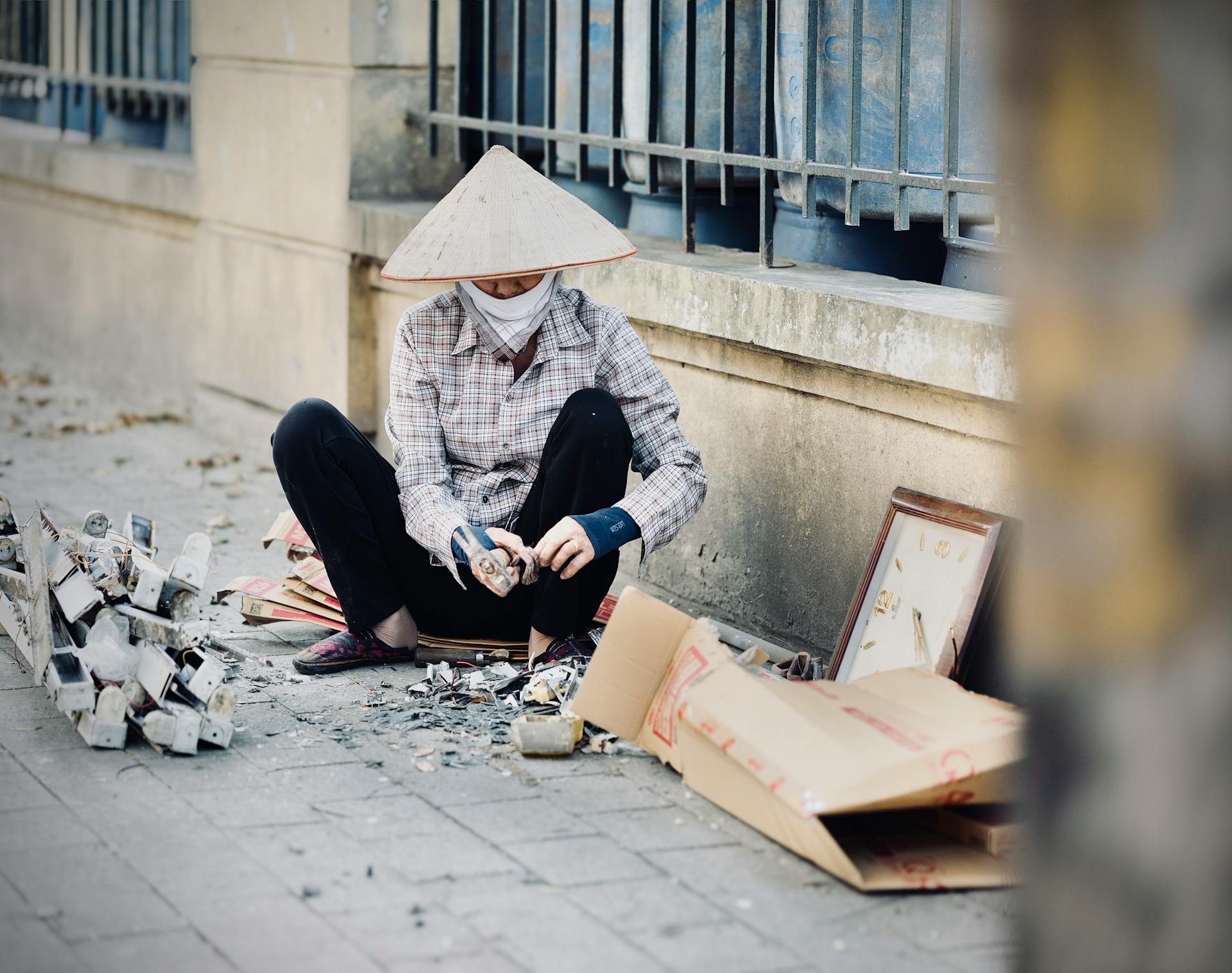Individual wearing conical hat sorting recyclables outdoors in urban setting.