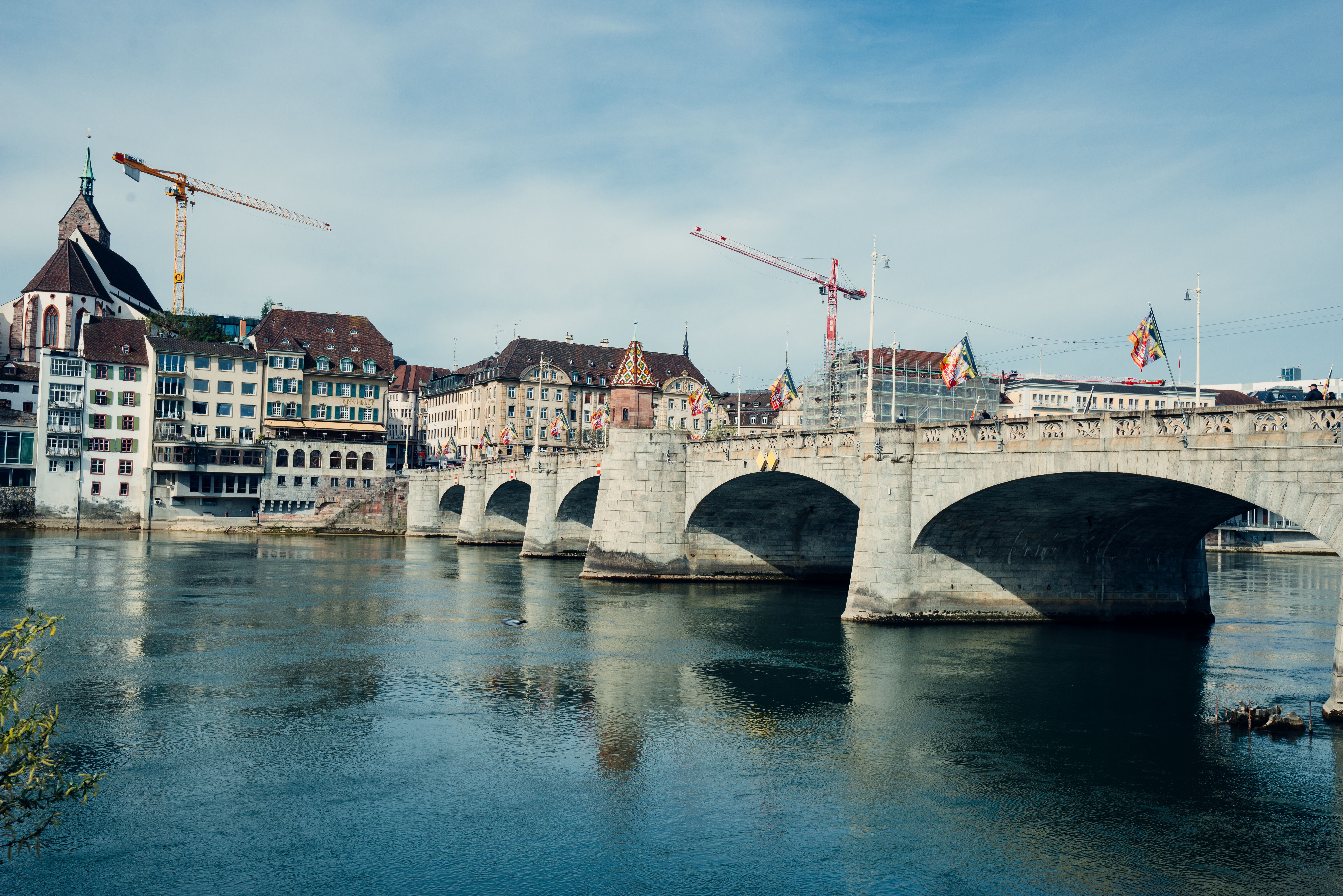 A picturesque scene of Basel's historic bridge with city buildings and cranes by the Rhine River.