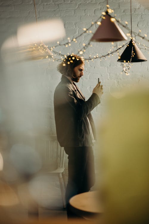 Fotografi Fokus Selektif Pria Berdiri Di Bawah Lampu Gantung Menggunakan Smartphone