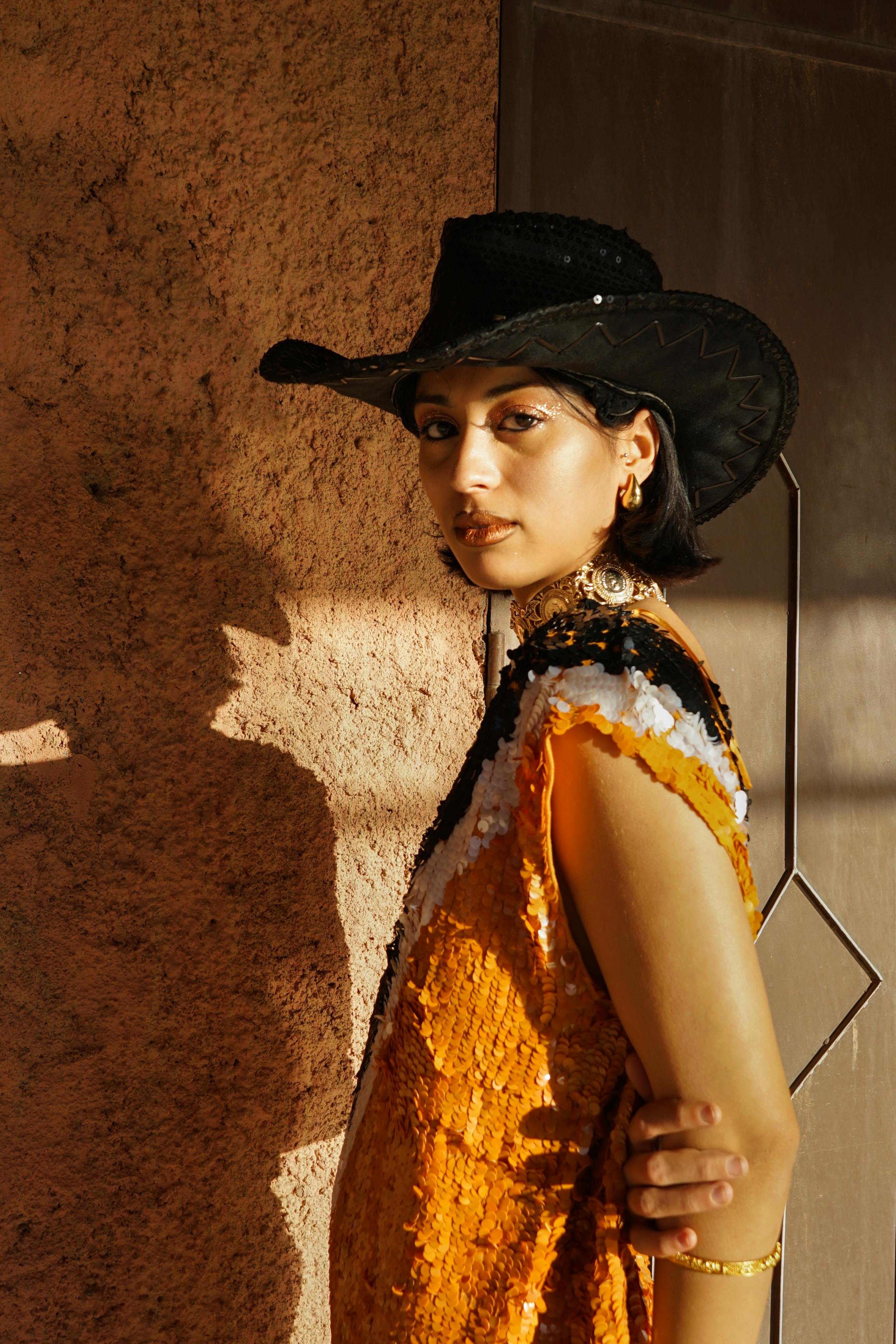 Stylish woman wearing a western hat poses against a textured wall in warm sunlight.