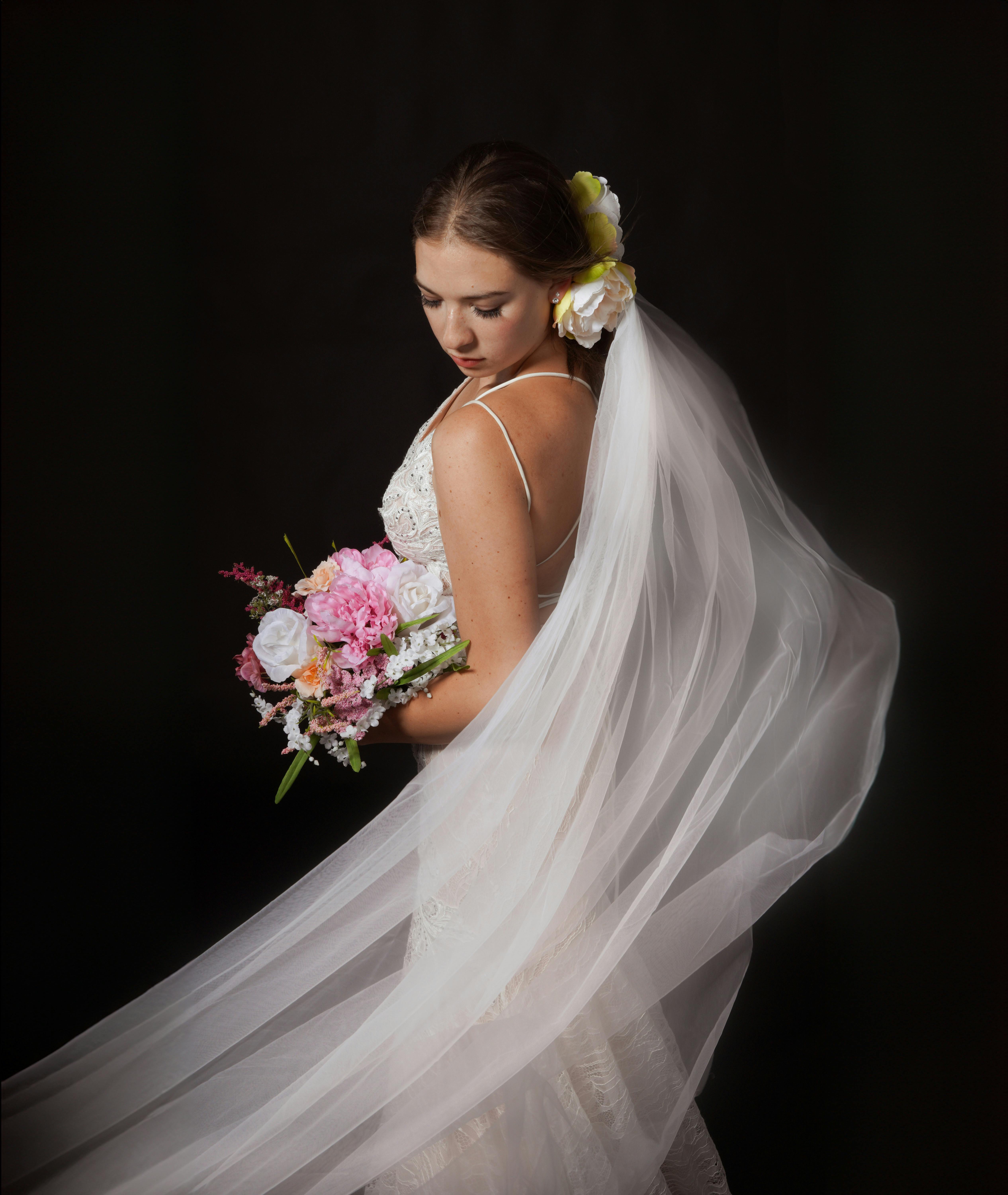Bride in white gown with flowing veil holding a bouquet against a dark backdrop.