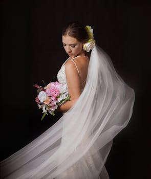 Bride in white gown with flowing veil holding a bouquet against a dark backdrop.