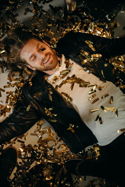 A joyful man in a leather jacket smiles amidst festive golden confetti, celebrating New Year's Eve.
