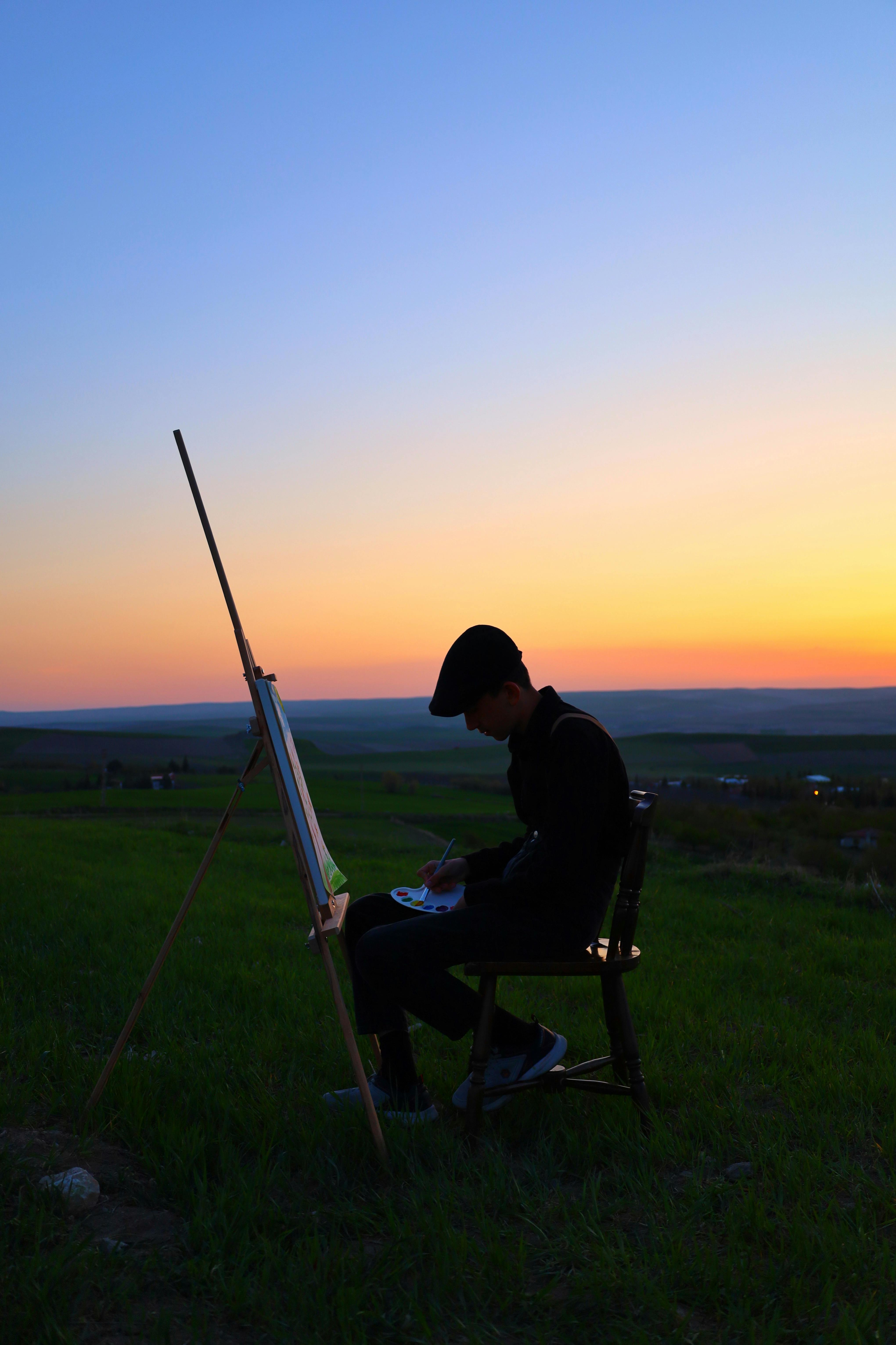 Silhouette of an artist painting on a canvas during sunset in a peaceful rural landscape.