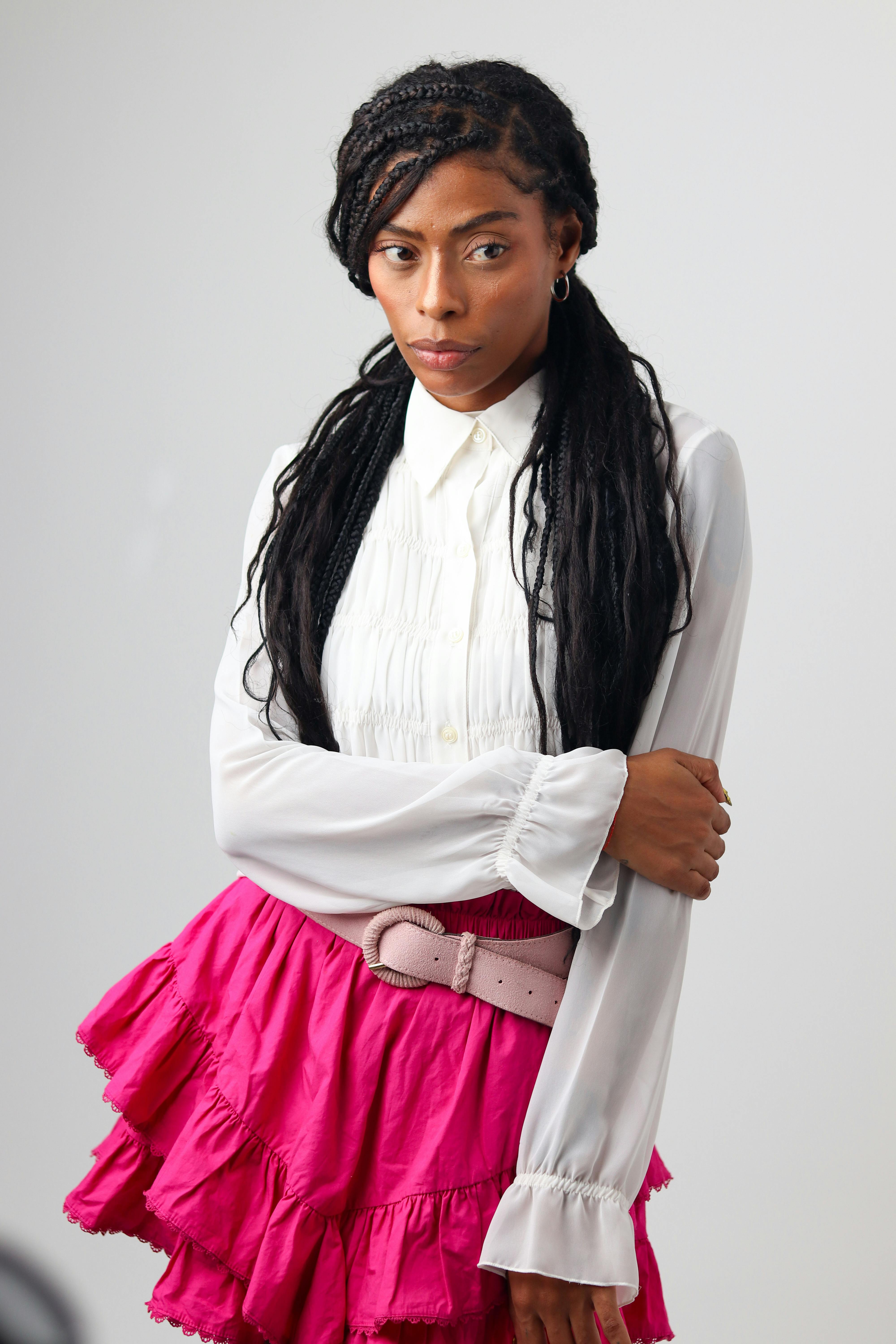 Gratis Una donna elegante con i capelli intrecciati posa con una gonna rosa acceso e una camicetta bianca su uno sfondo neutro. Foto a disposizione