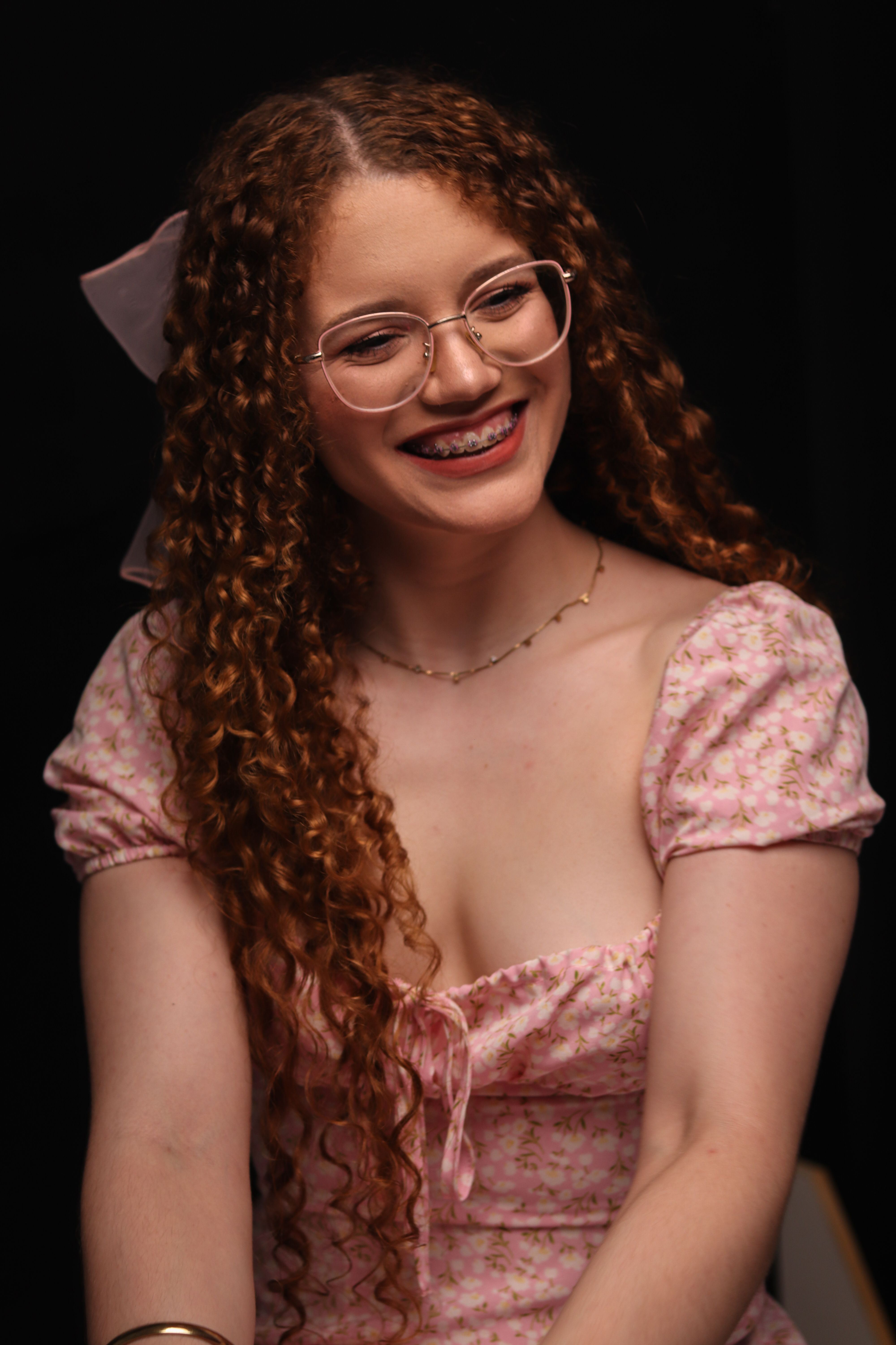 A joyful young woman with curly hair, glasses, and a floral dress, captured indoors.