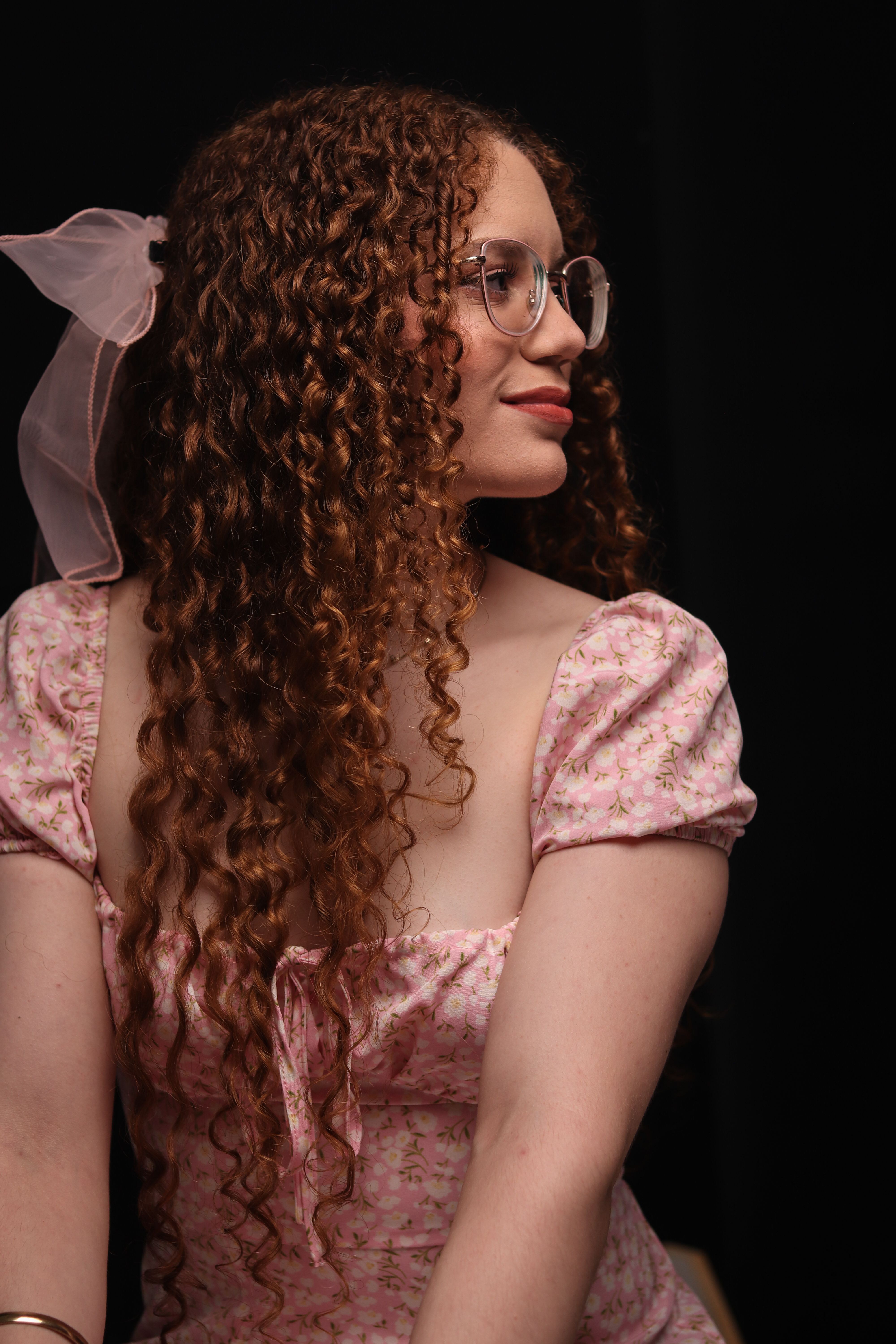 A young woman with curly hair and glasses wearing a pink floral dress poses against a dark background.