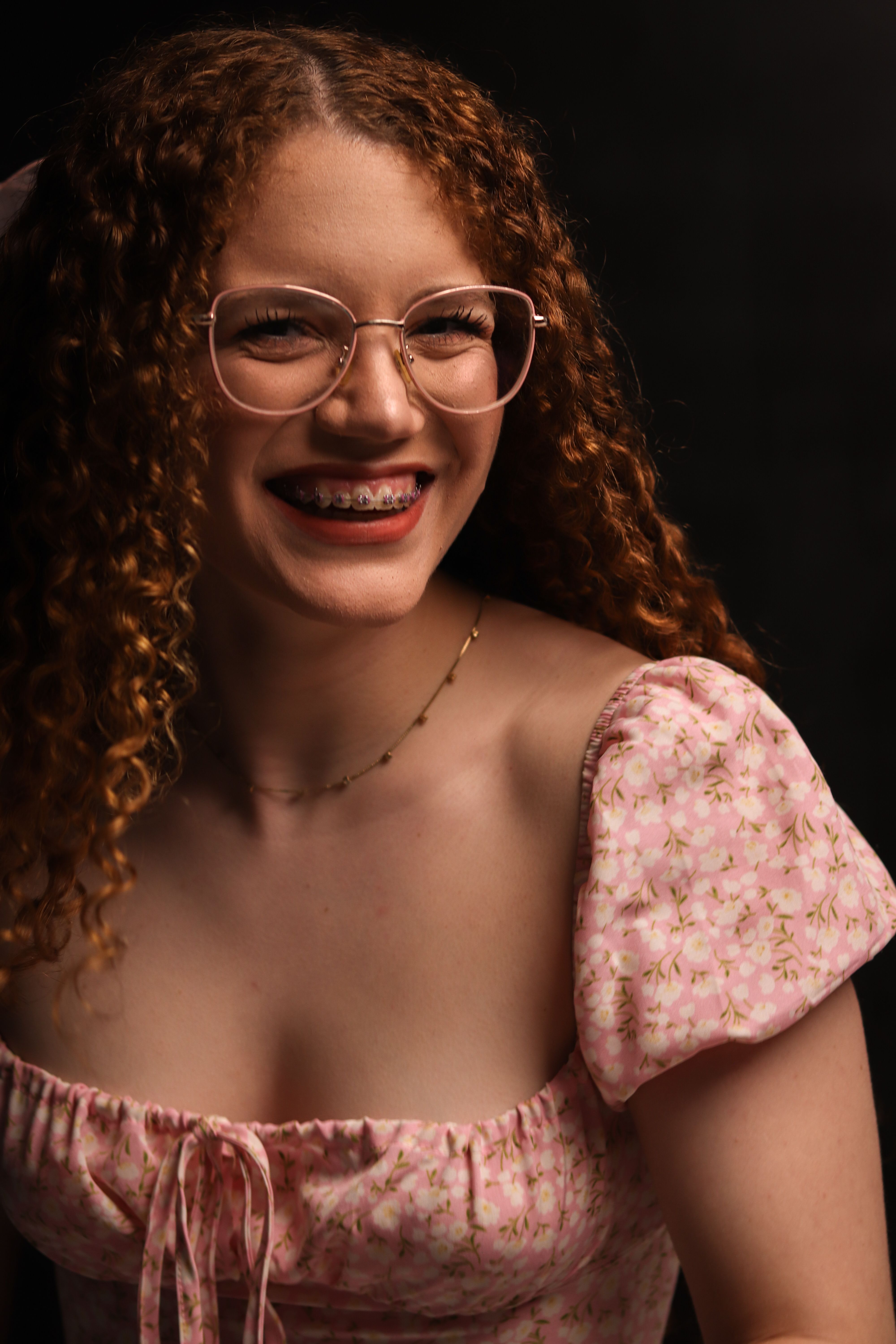 Smiling woman with red curly hair, glasses, and braces in pink floral dress.