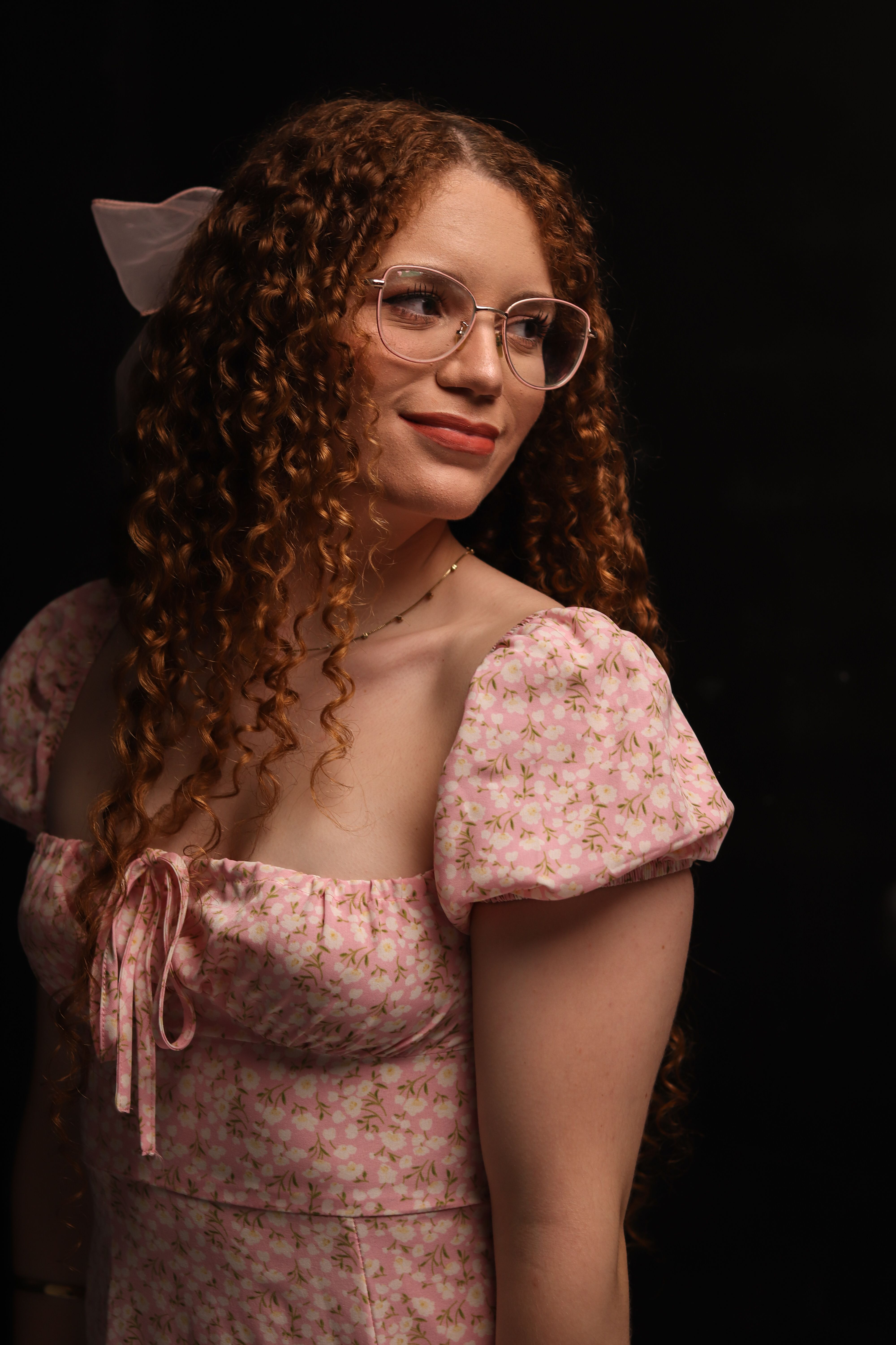 A beautiful woman with curly hair and glasses wearing a pink floral dress, looking to the side with a serene expression.