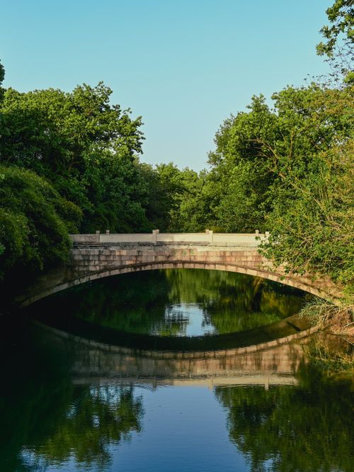 Free Peaceful stone bridge over a calm river with lush green surroundings under a clear sky. Stock Photo