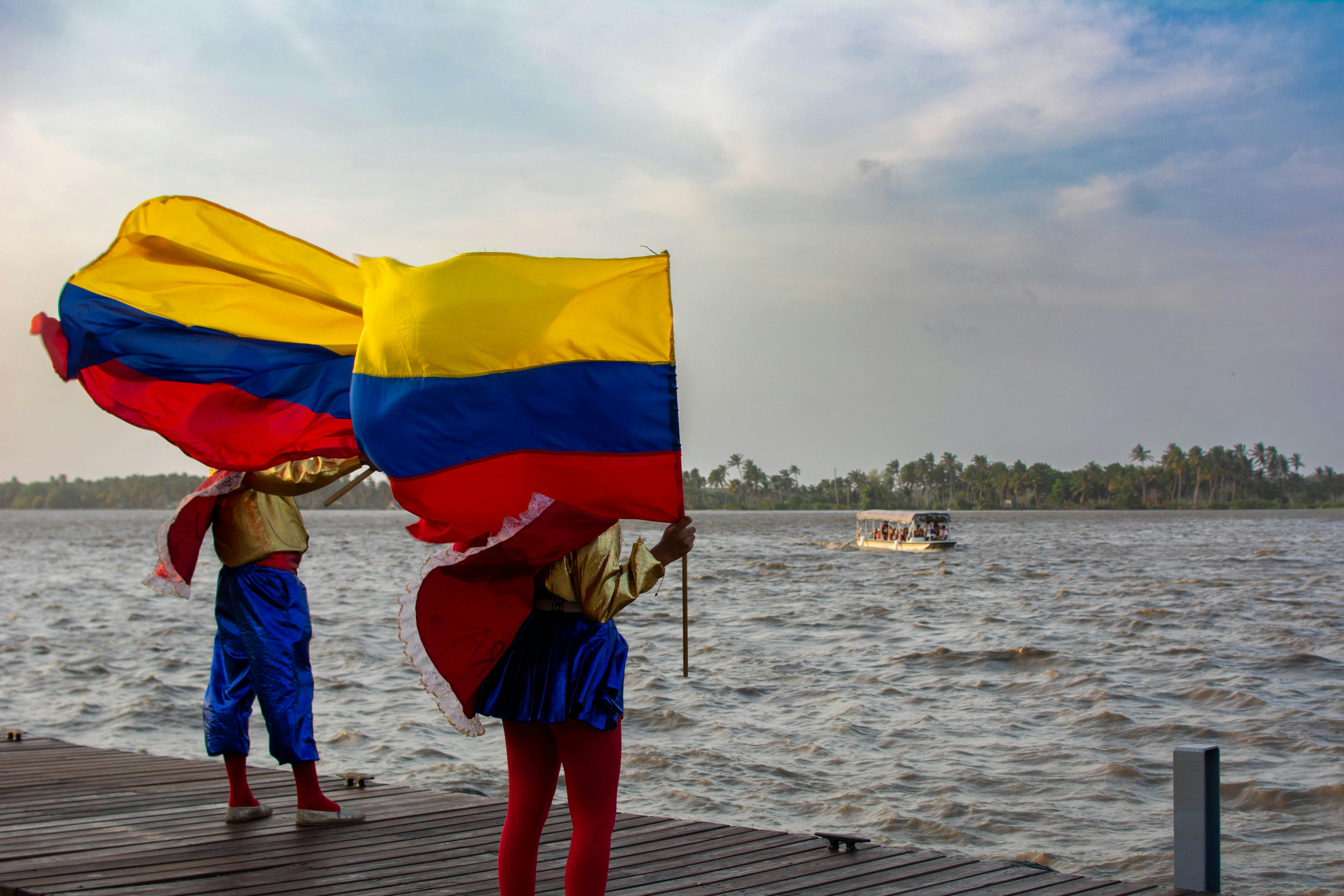 Gratis Banderas colombianas vibrantes ondeando sobre un río pintoresco con trajes coloridos. Foto de stock