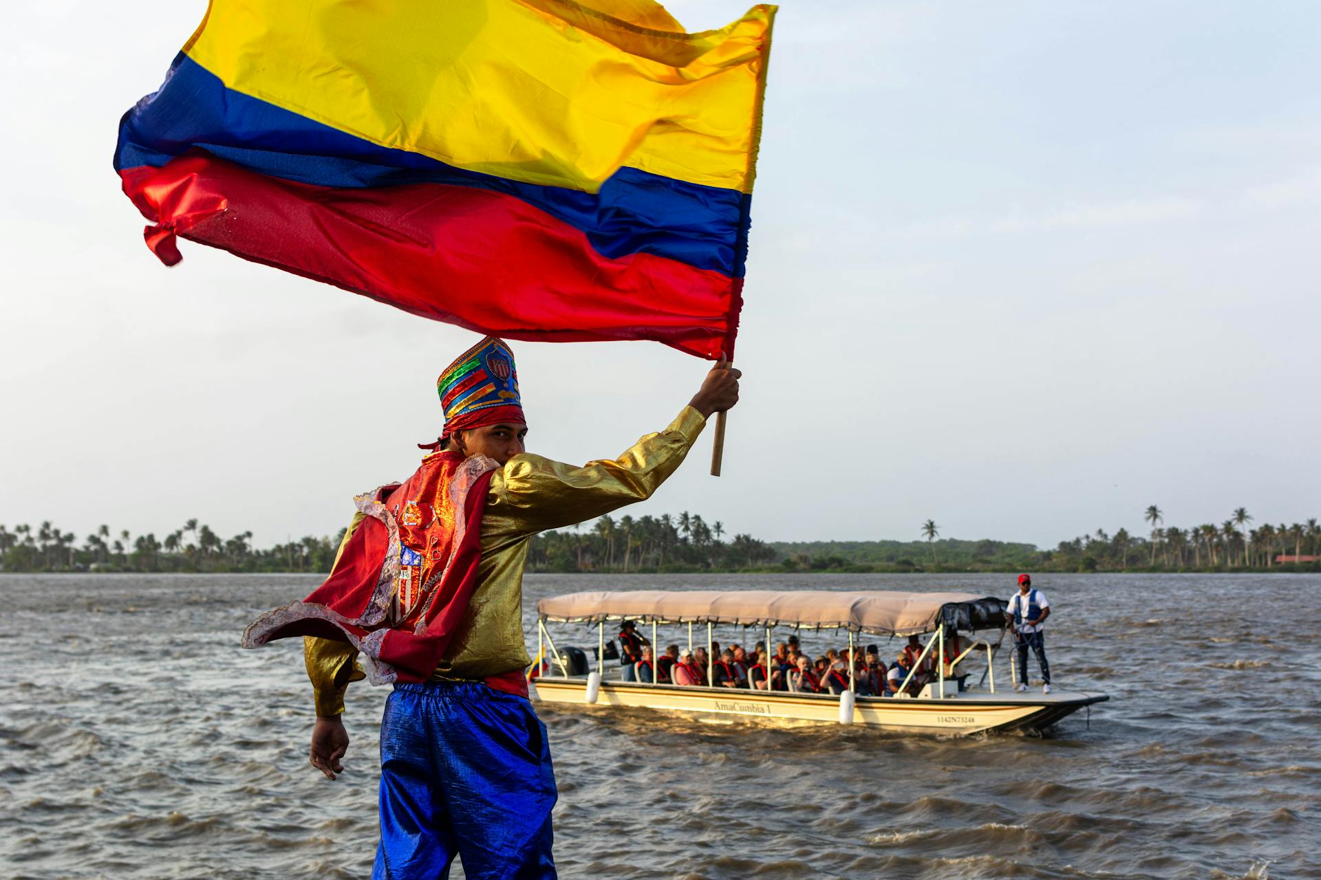 A vibrant parade performer waves the Colombian flag next to a tourist boat on the river.