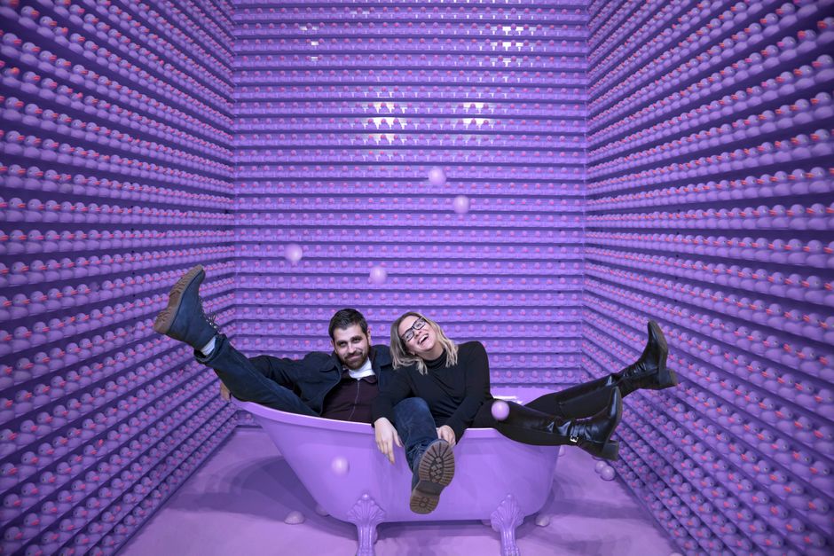 Two adults enjoying a whimsical moment in a bathtub surrounded by purple ducks.