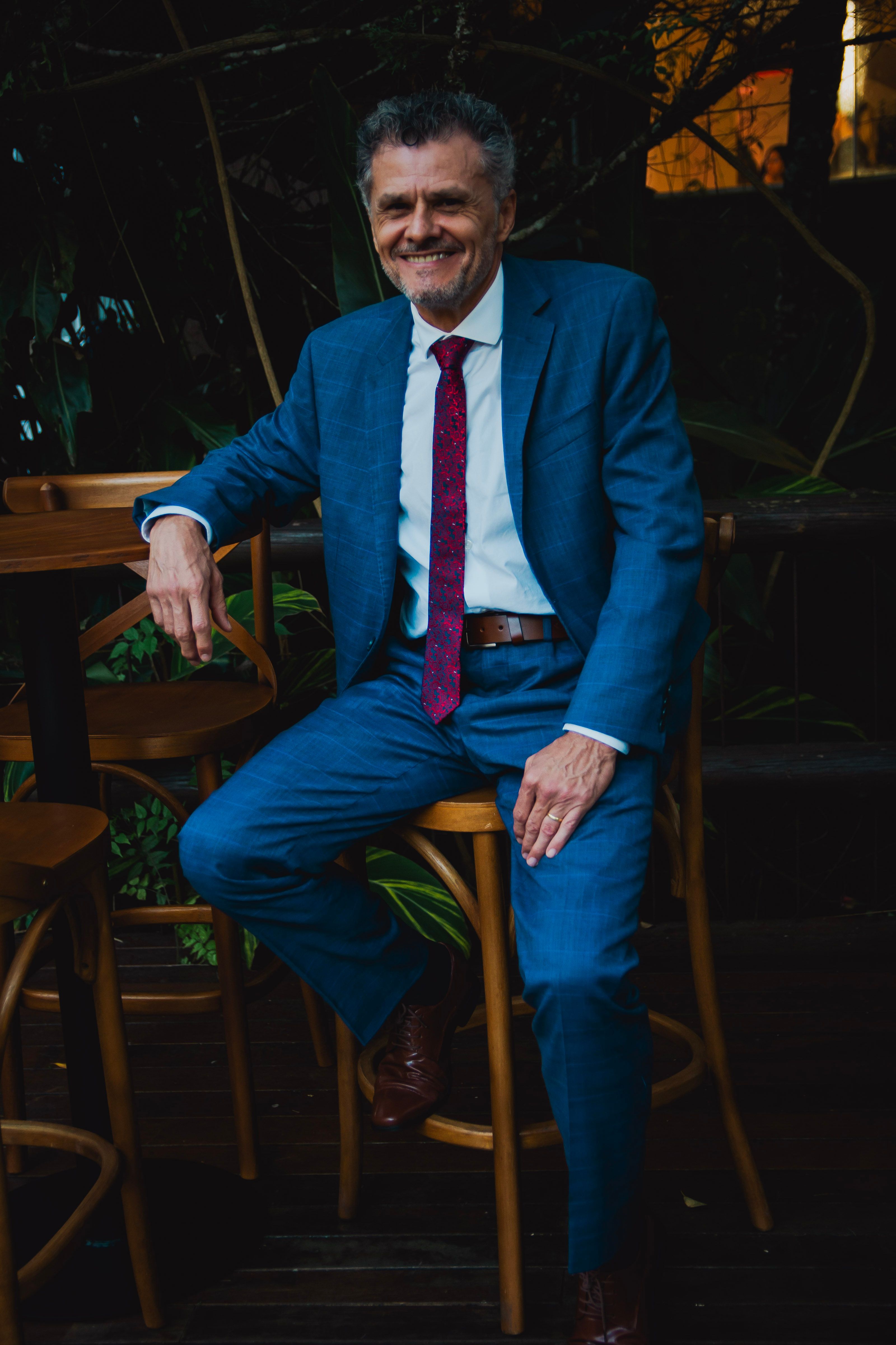 A smiling man in a blue suit and red tie sits casually outdoors on a wooden chair.