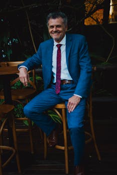 A smiling man in a blue suit and red tie sits casually outdoors on a wooden chair.