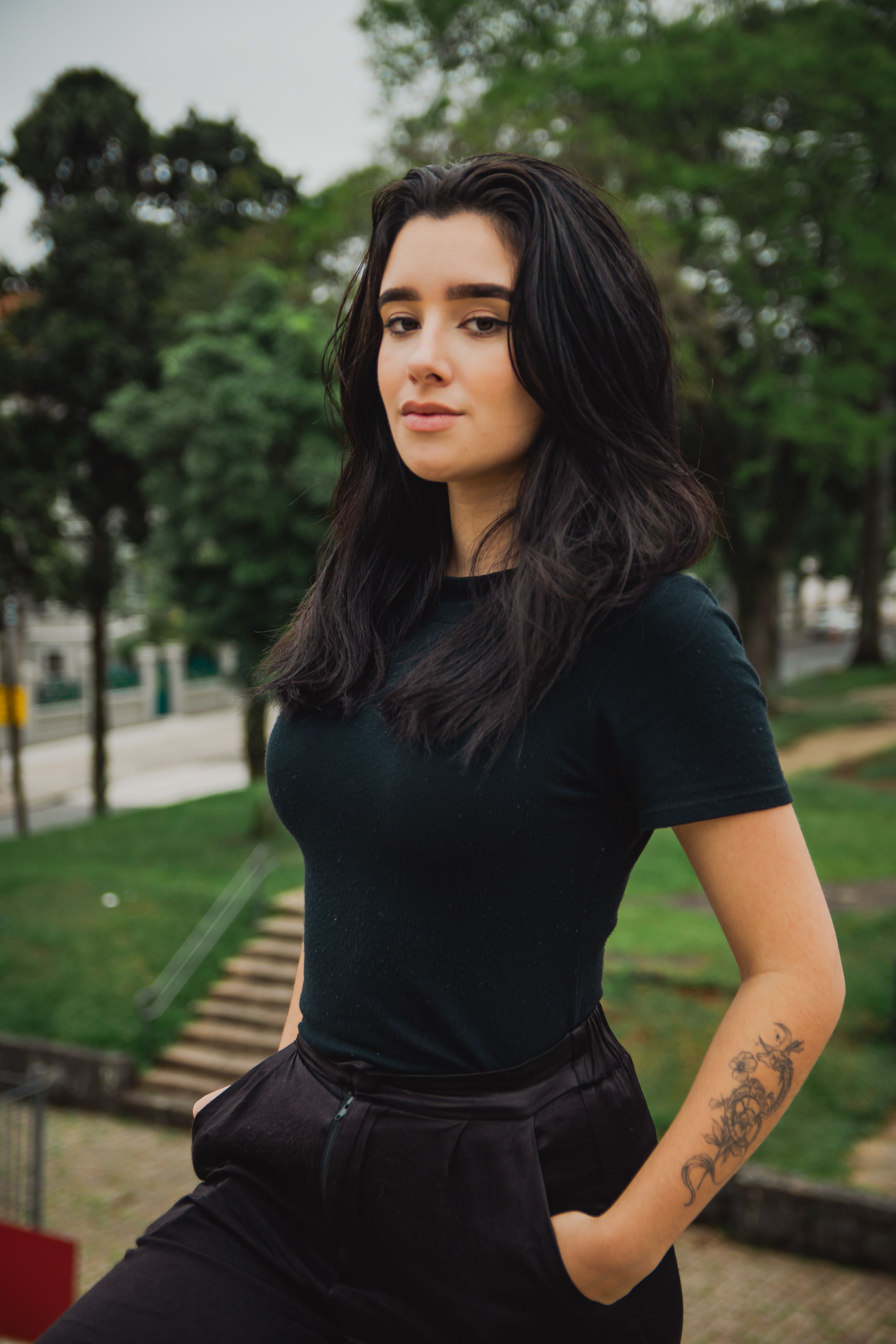 Confident woman with long black hair posing outdoors in a green urban park.
