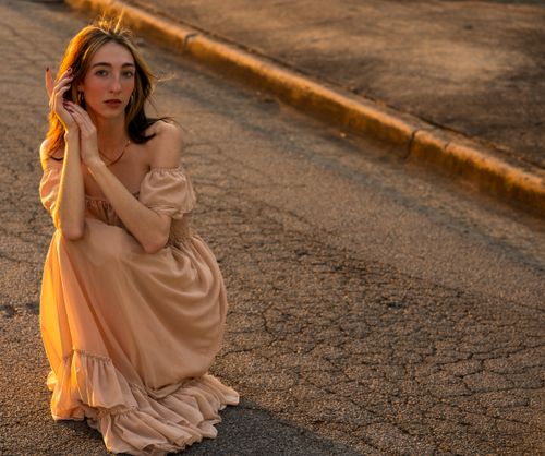 Free A woman in a flowing dress sits on a street, illuminated by warm sunset light. Stock Photo