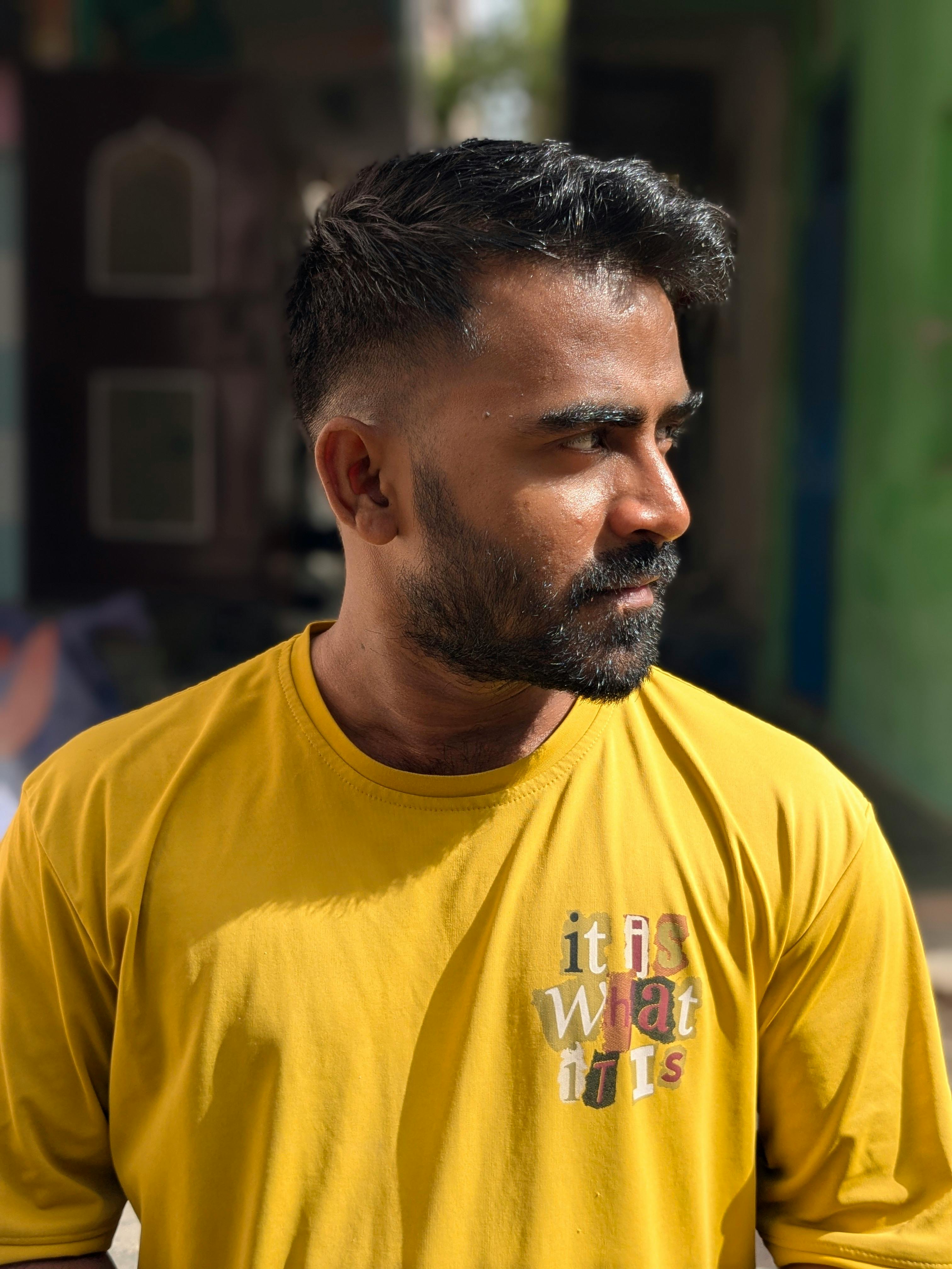 Portrait of a young man in a bright yellow shirt with 'it is what it is' text, looking pensive outdoors.