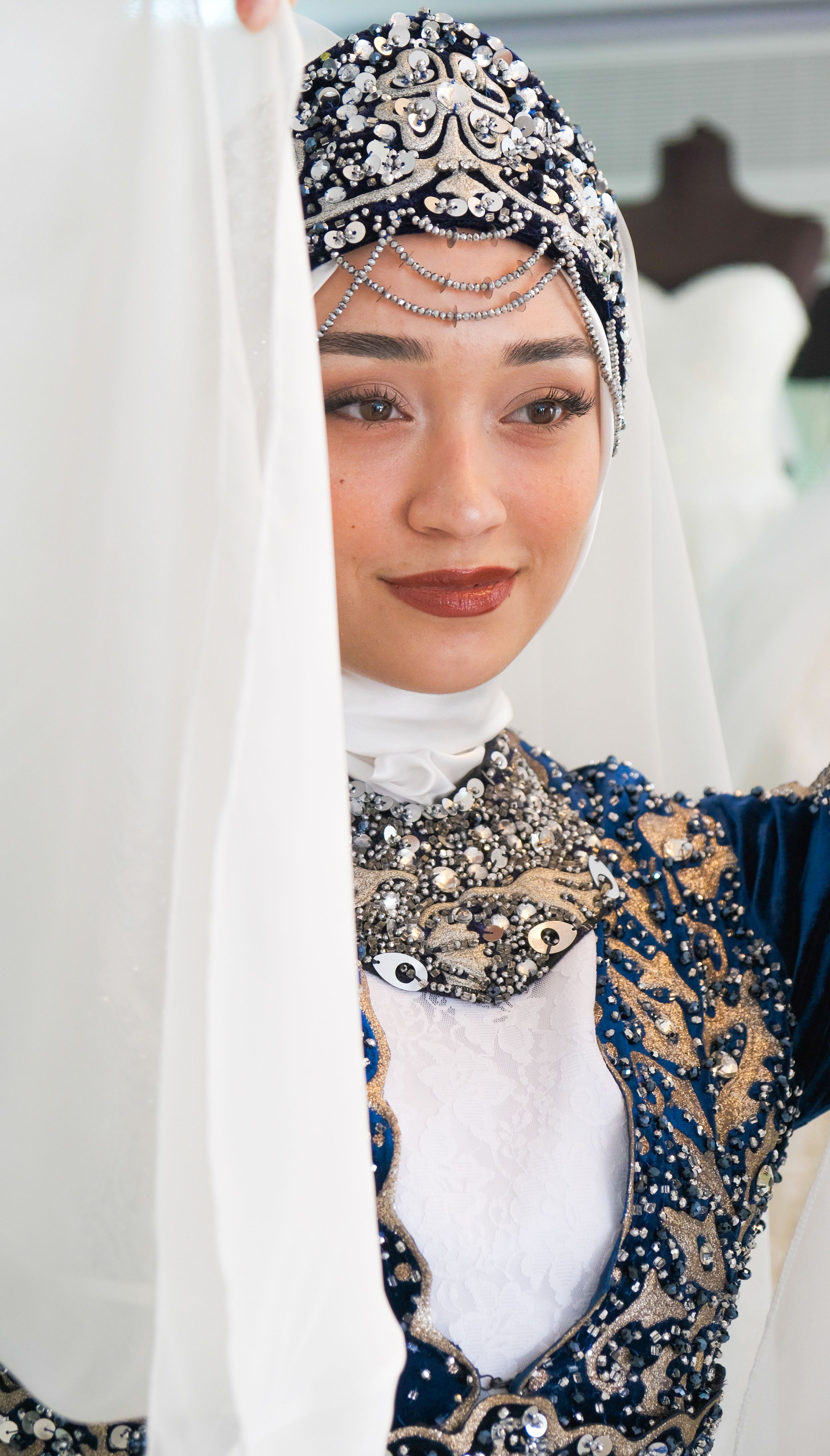 A woman in detailed traditional dress with jewel embellishments poses gracefully indoors.