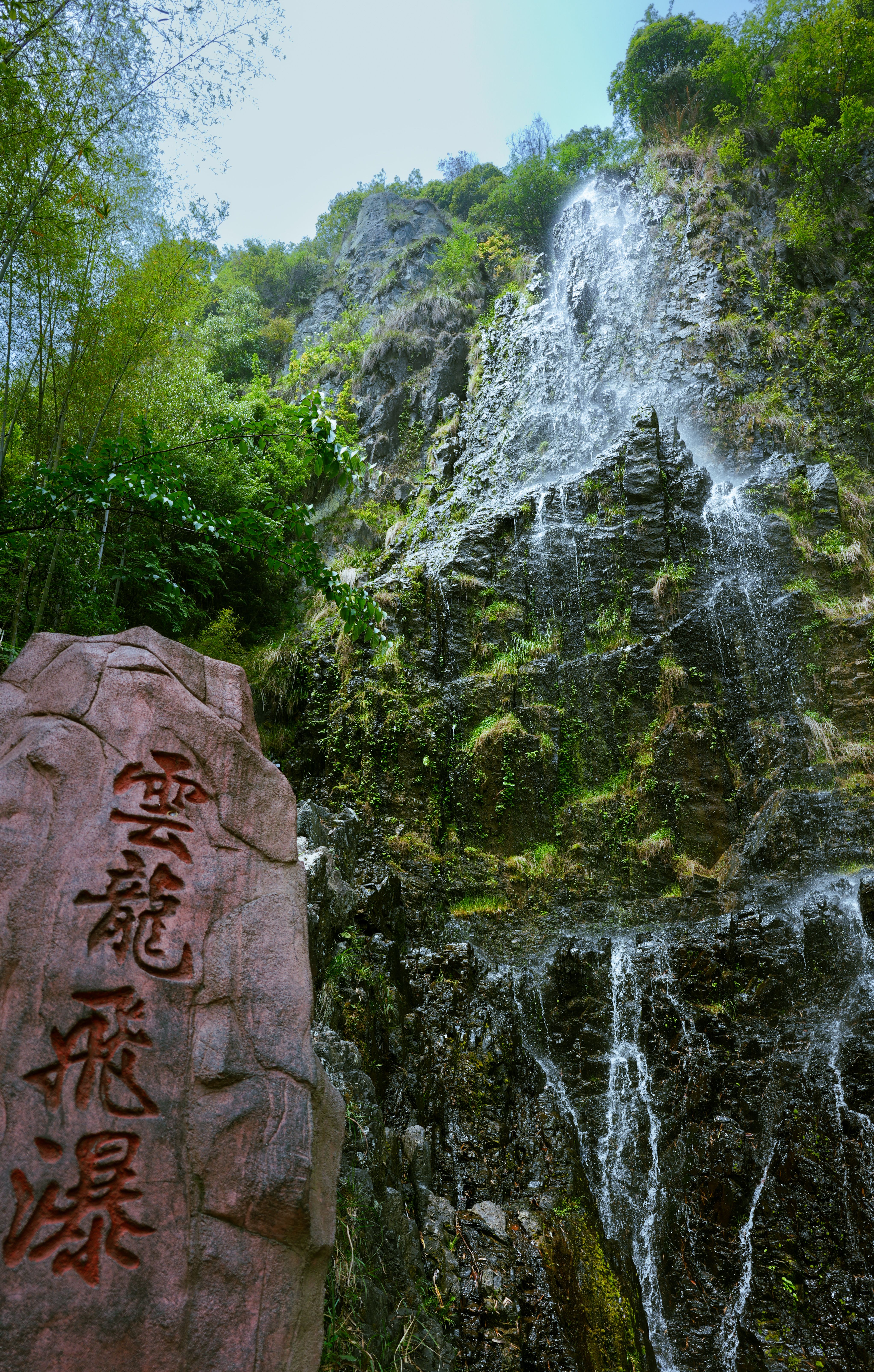 Stunning waterfall cascading down rocky cliff with stone featuring Chinese characters, surrounded by lush greenery.