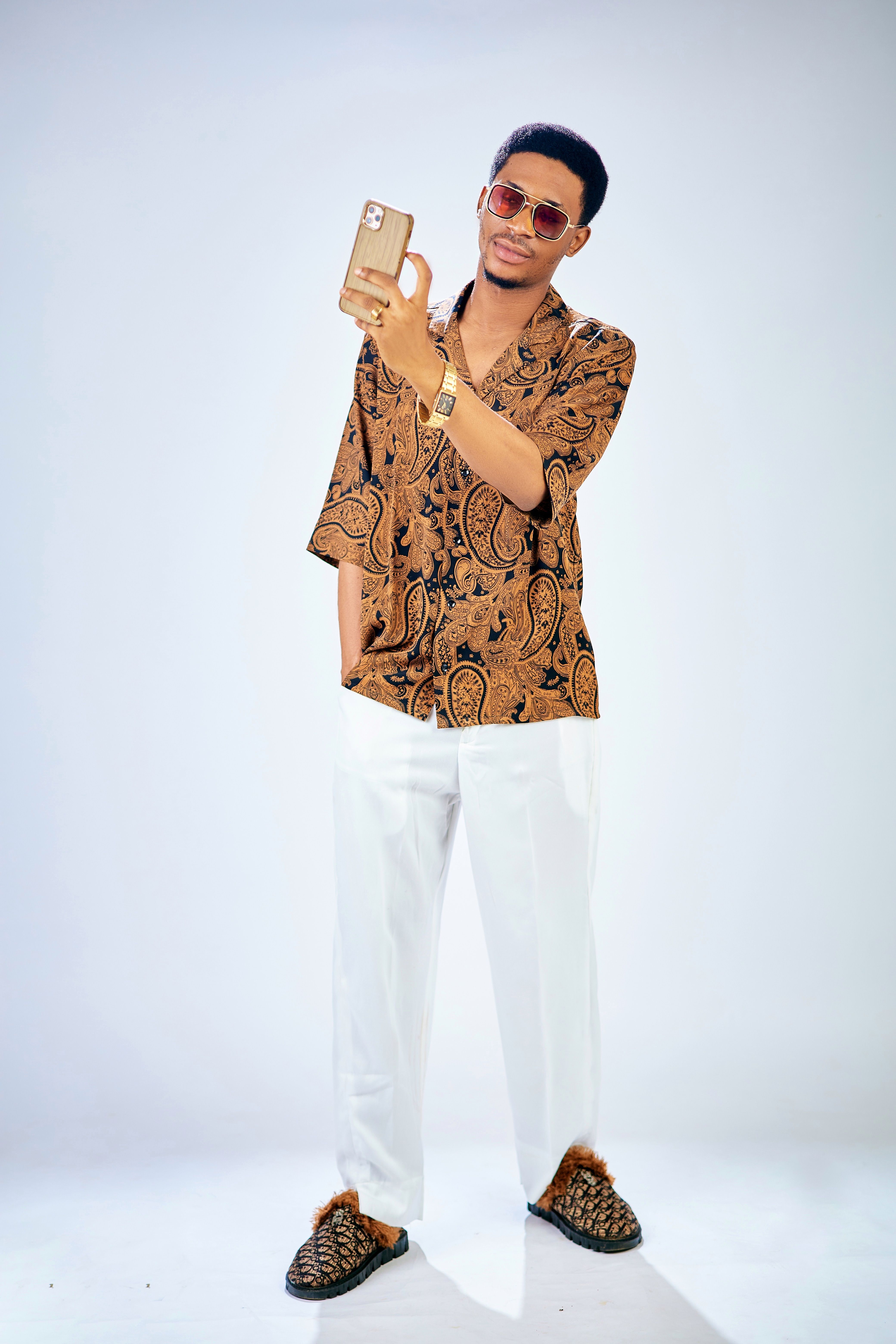Trendy man in sunglasses taking a selfie wearing a patterned shirt and white pants indoors.