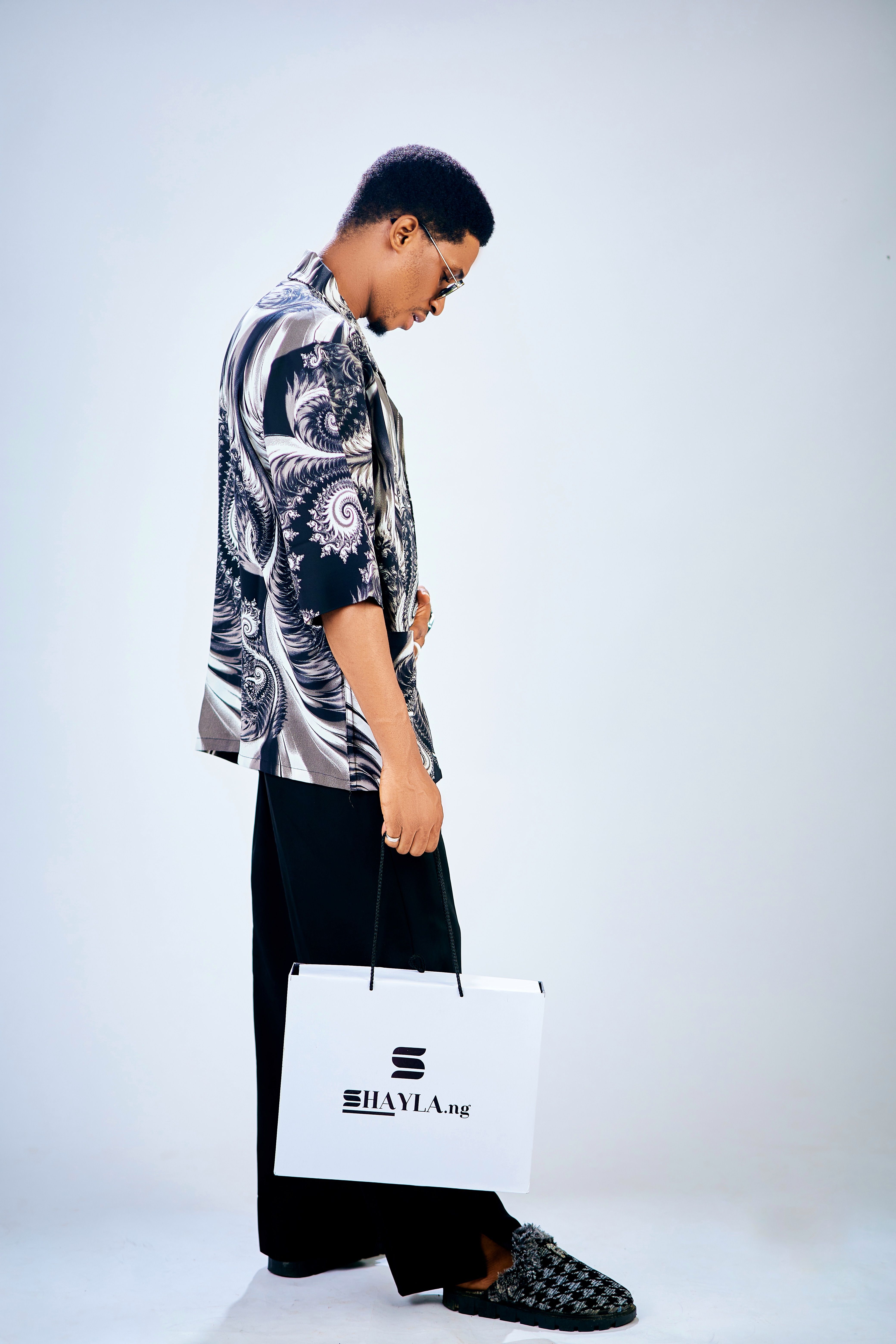 Stylish person in a studio holding a branded shopping bag, wearing patterned clothing.