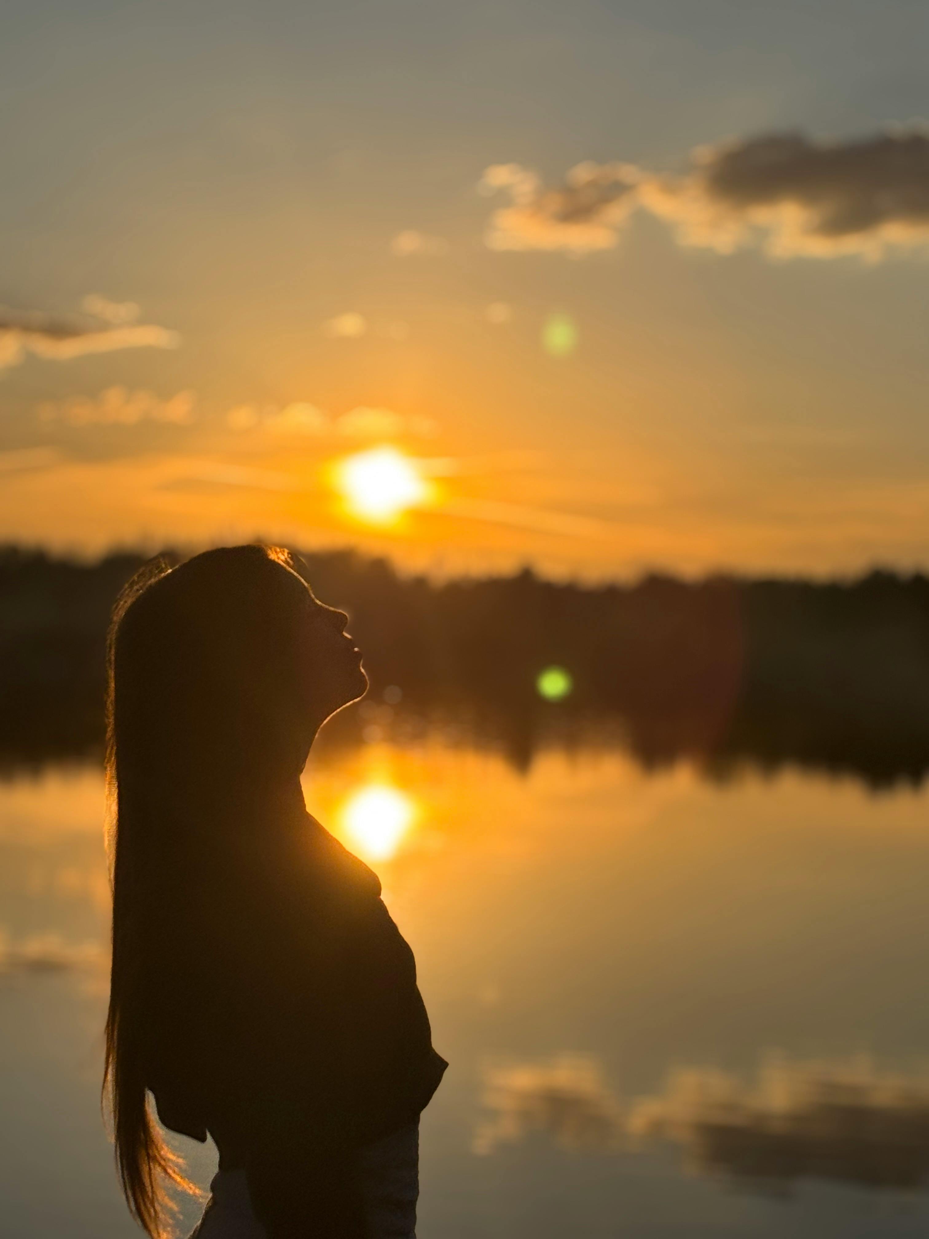 Silhouette of a woman enjoying sunset by a tranquil lake reflecting the sky.