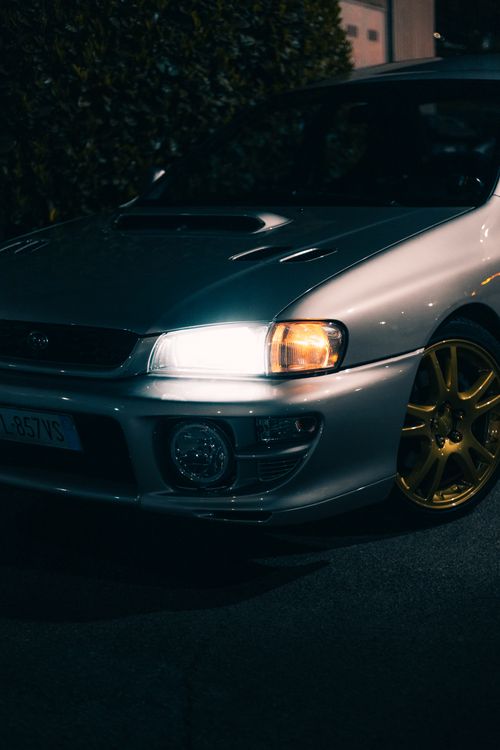 Free A stylish silver car with a headlight on and golden wheels, parked outdoors at night. Stock Photo