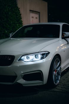 White sports car with illuminated headlights parked outdoors at night.
