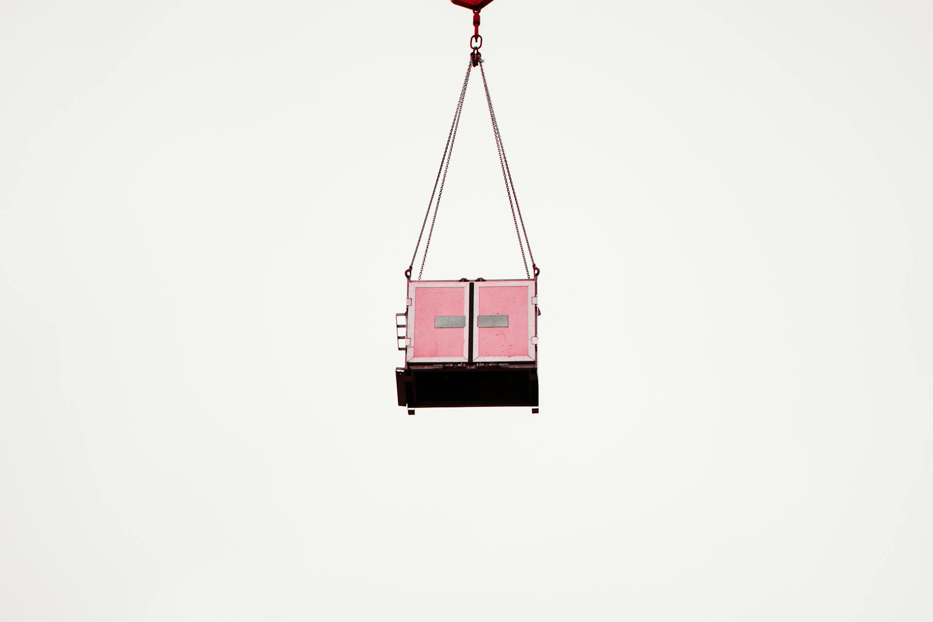 A pink cargo container suspended mid-air against a clear white sky, minimalist composition.