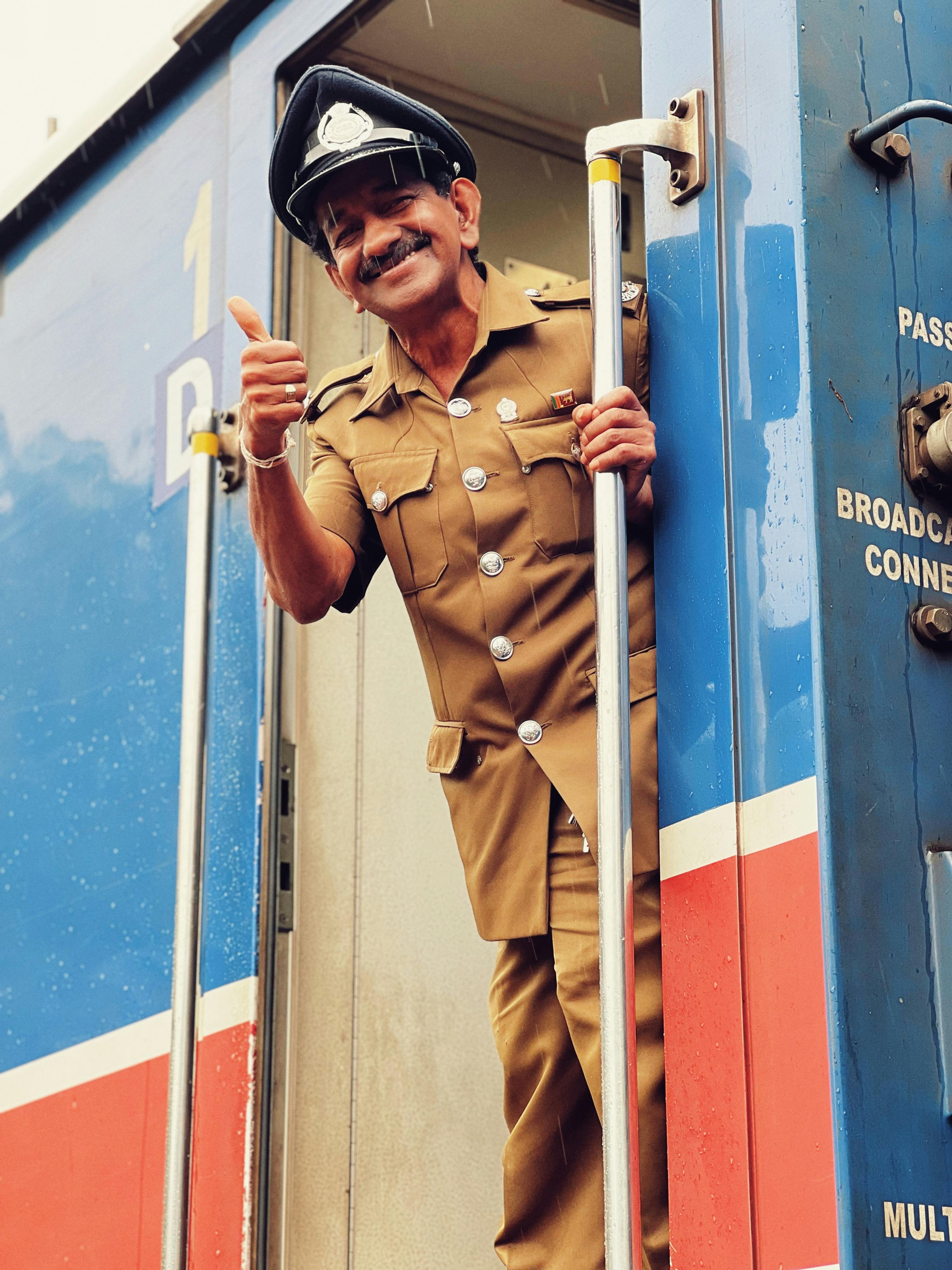 A cheerful Sri Lankan train conductor in uniform giving a thumbs up from the train entrance.