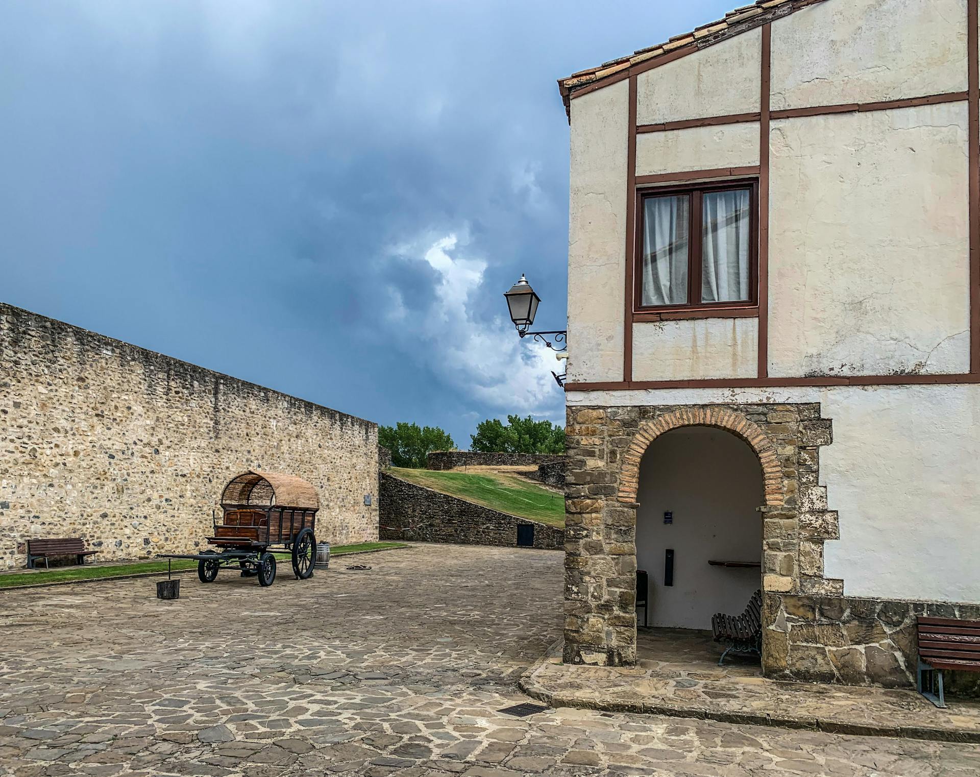 A rustic courtyard scene featuring a historic wagon and stone architecture under dramatic stormy skies.