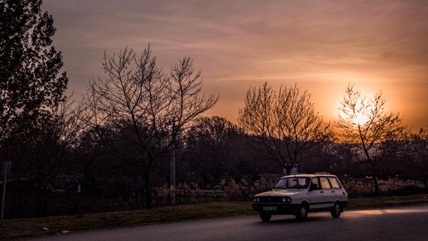 A vintage car traveling on a scenic road during a beautiful sunset, framed by leafless trees.